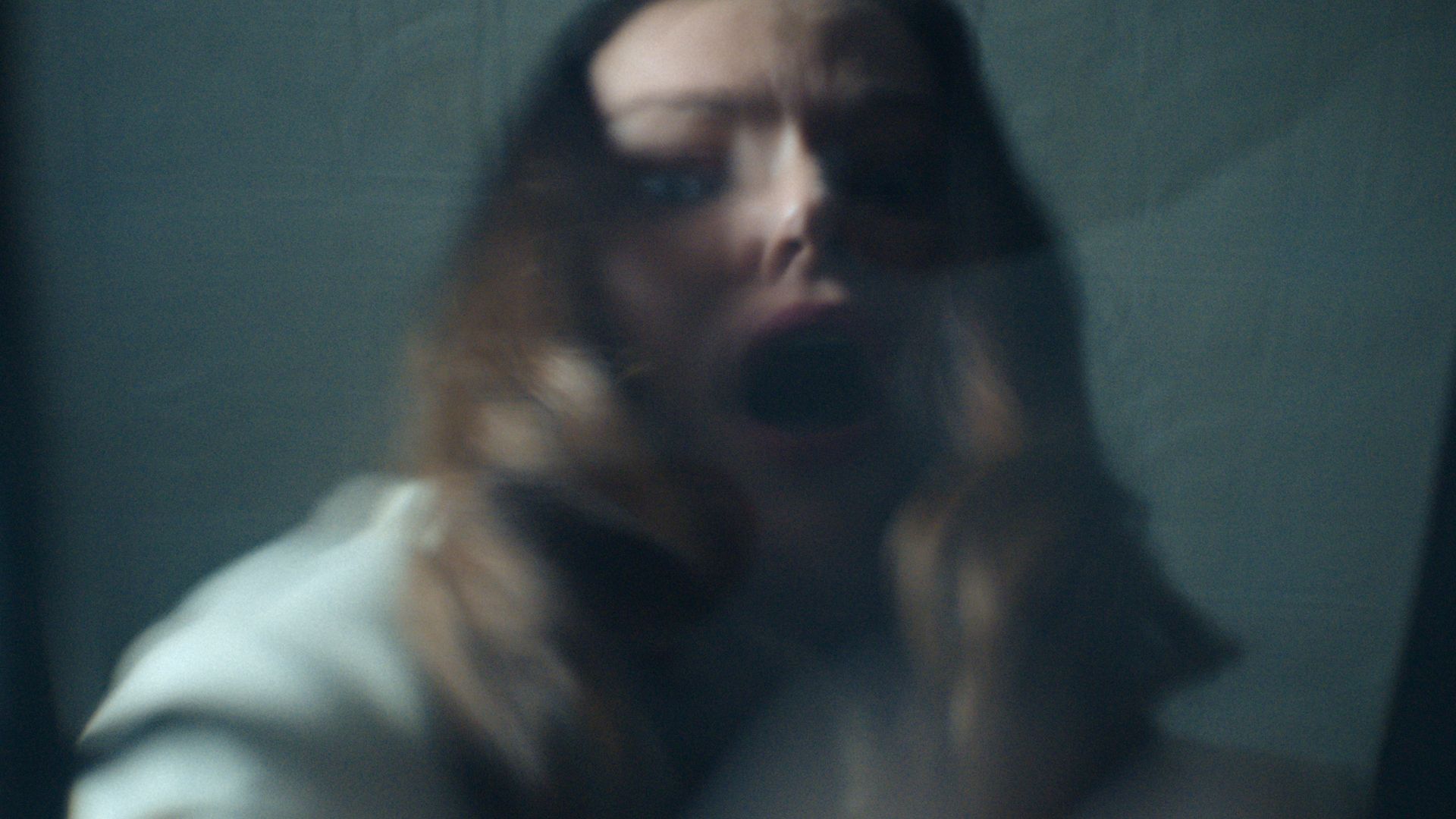 Blurry close-up of a woman with long hair, mouth open wide, and a distressed expression, set against a dark, muted background.