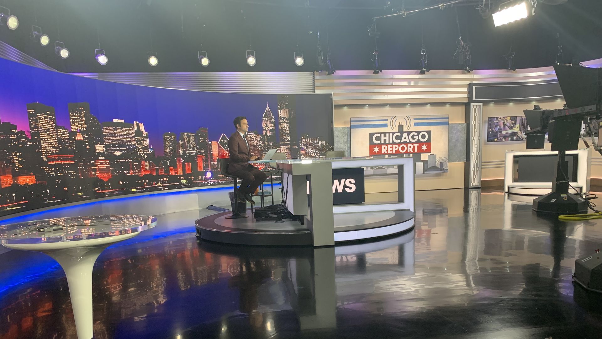 News studio set with a man in a suit at a desk with a laptop, a Chicago skyline backdrop, and a bold "CHICAGO REPORT" sign. Studio lights and camera rigs visible.