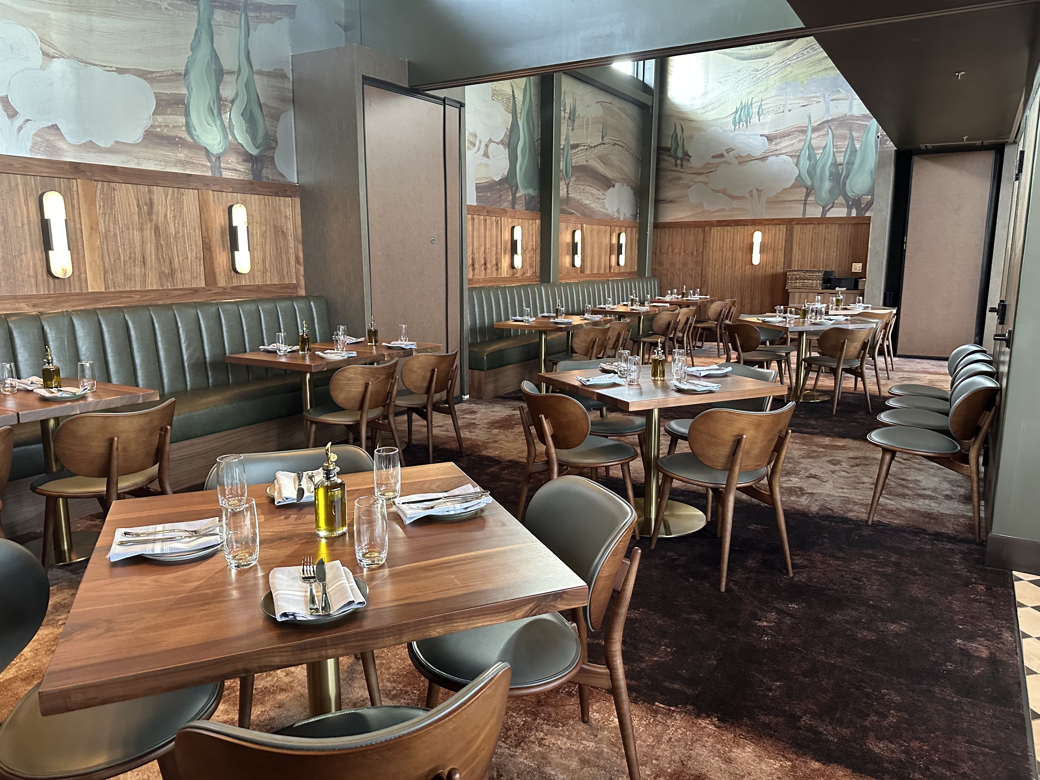 Interior of a stylish restaurant with wood paneling, green cushioned banquettes, and many wooden tables set with napkins, glasses, and olive oil bottles. A mural of trees and clouds along the wall.