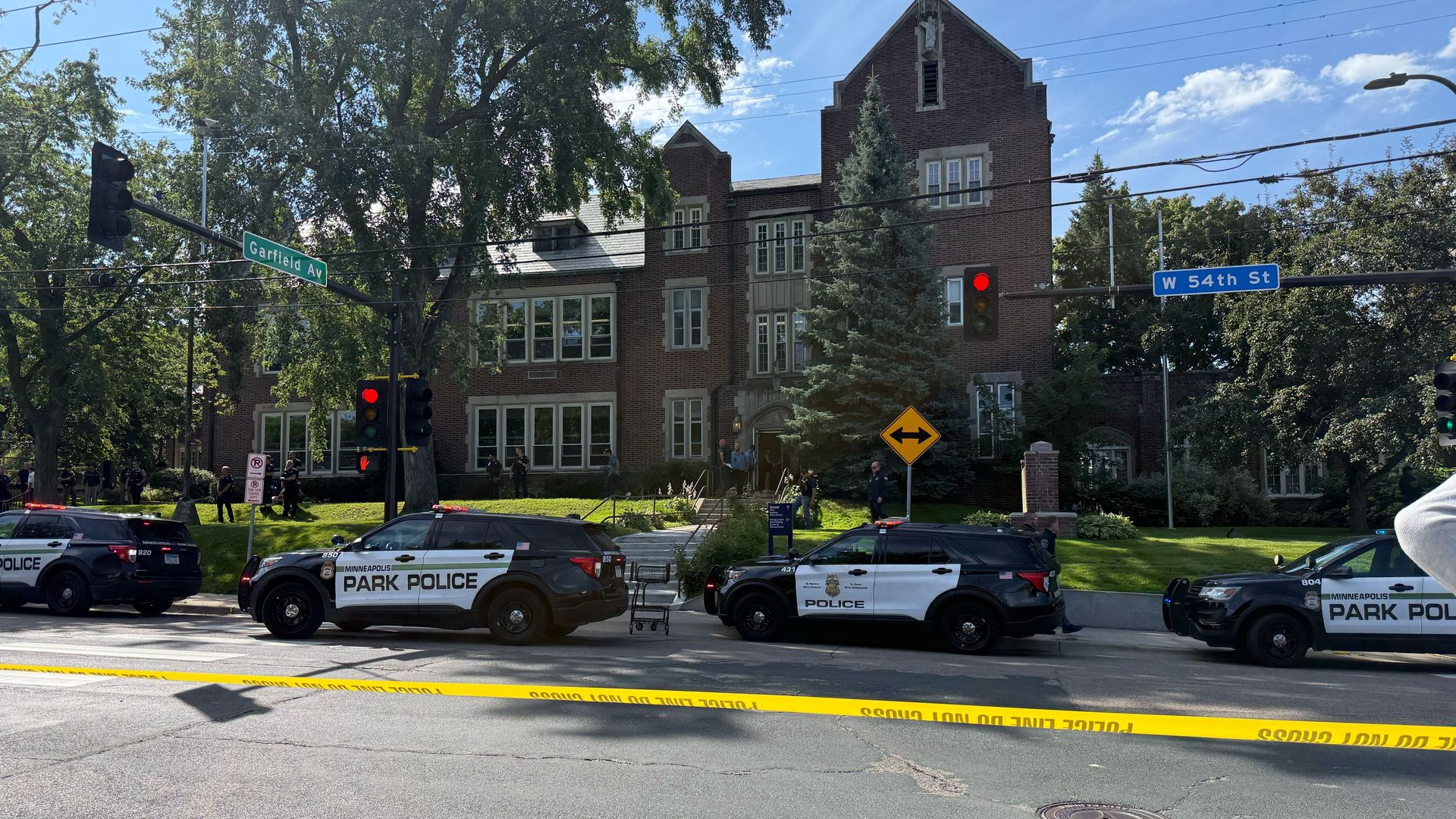 Four black and white Minneapolis Park Police SUVs block a street corner at Garfield Av and W 54th St, with yellow police tape across the road in front of a large brick building and trees.