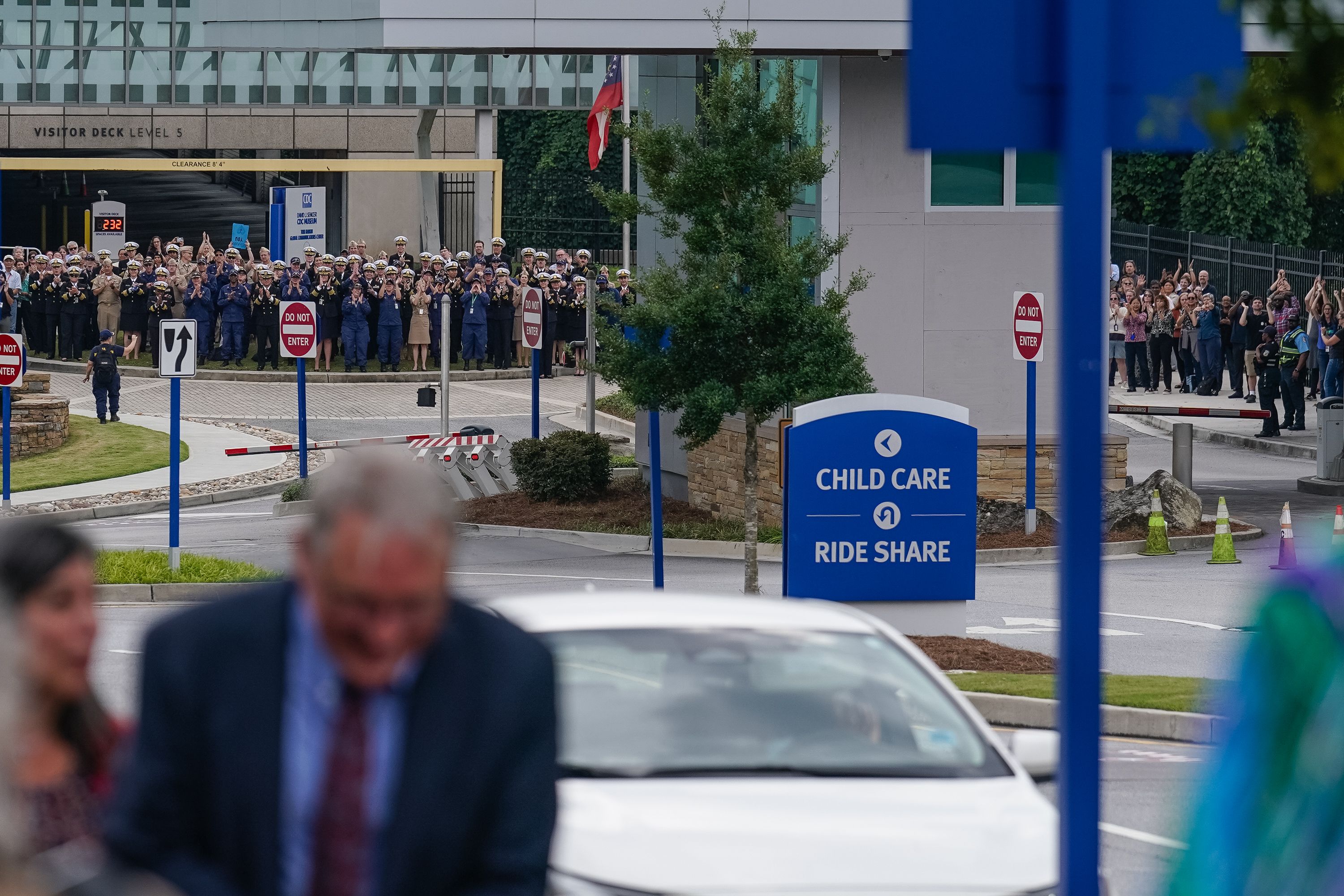 A group of uniformed personnel and other people stand and clap near a Visitor Deck sign, with a blue sign indicating directions to Child Care and Ride Share in the foreground.