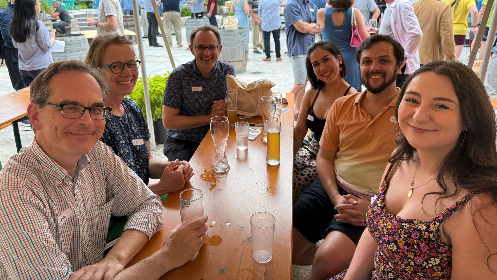 Six people smiling at a wooden table with drinks at an outdoor social event, with other groups mingling near a white building and greenery in the background.