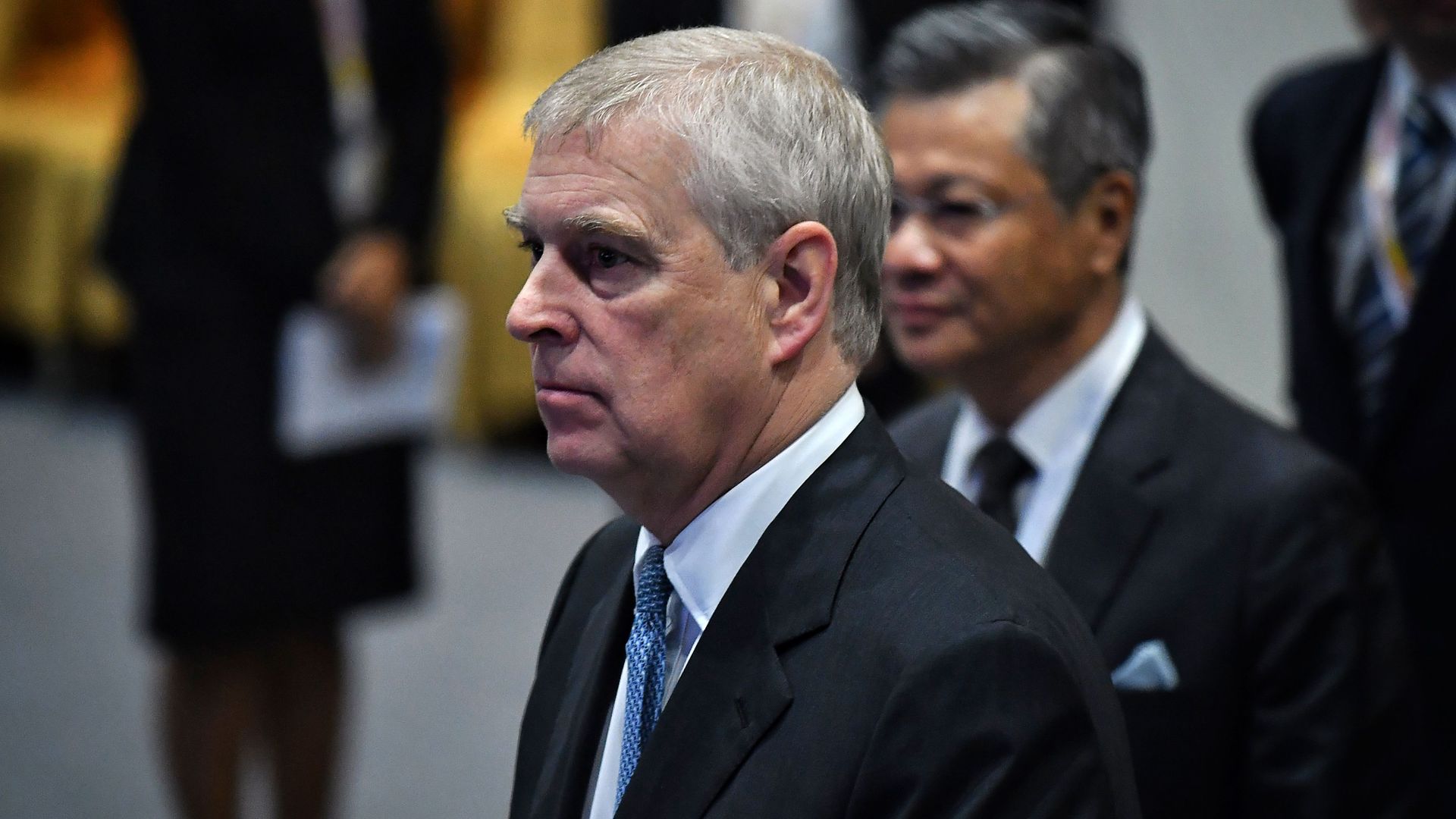 In this image, Prince Andrew stands and walks while wearing a suit.