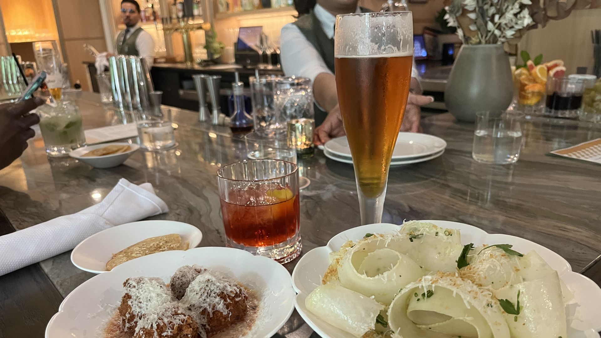Two white dishes on a marble bar counter: one with three meatballs topped with grated cheese, another with thinly shaved white vegetable curls garnished with herbs and cheese. Two drinks, amber beer in a tall glass and a red cocktail with an orange slice, are behind the dishes.