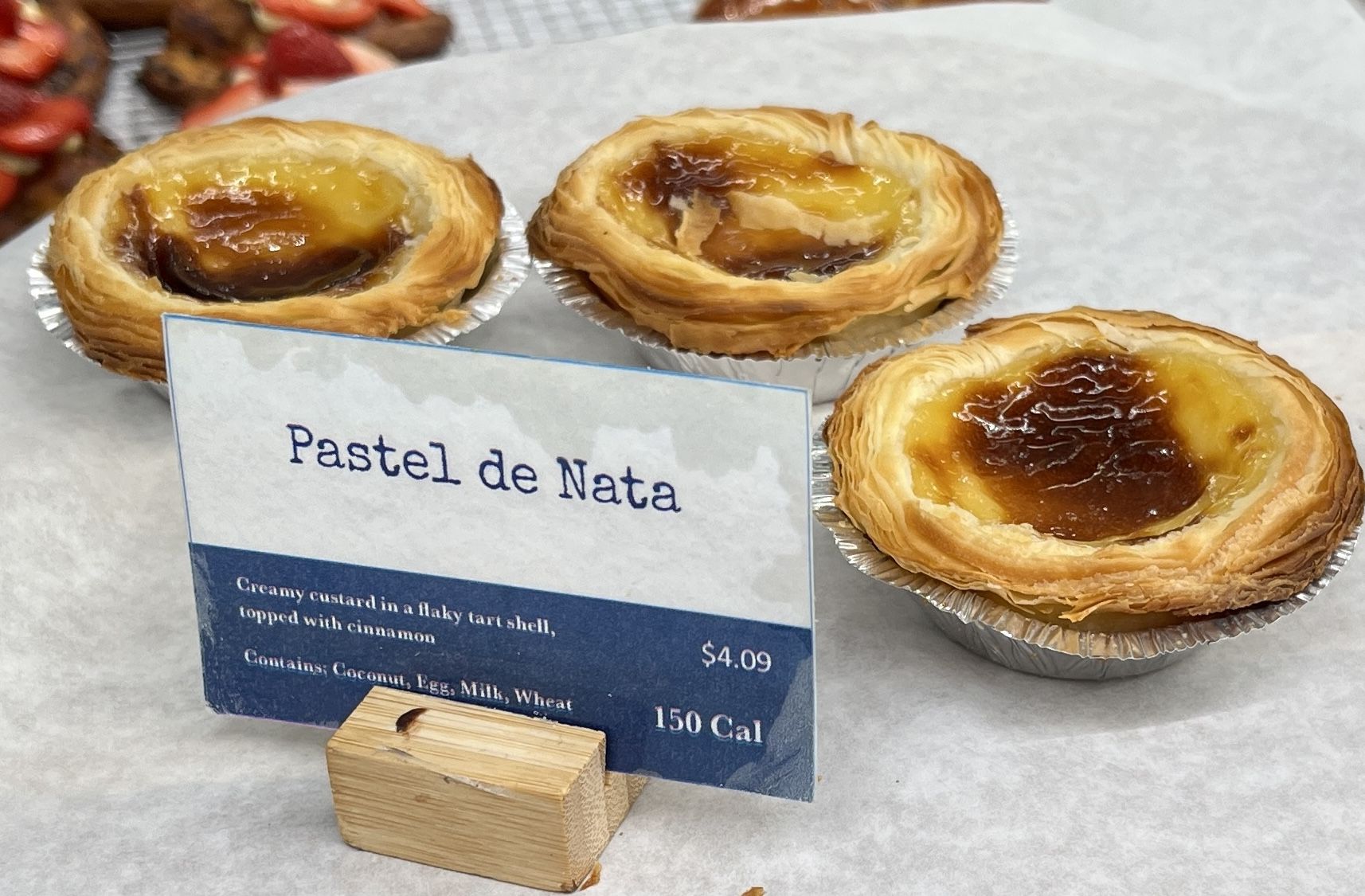 Three golden-brown Pastel de Nata Portuguese custard tarts in foil cups on a white surface, with a sign describing them as creamy custard in flaky shell, topped with cinnamon, priced $4.09.