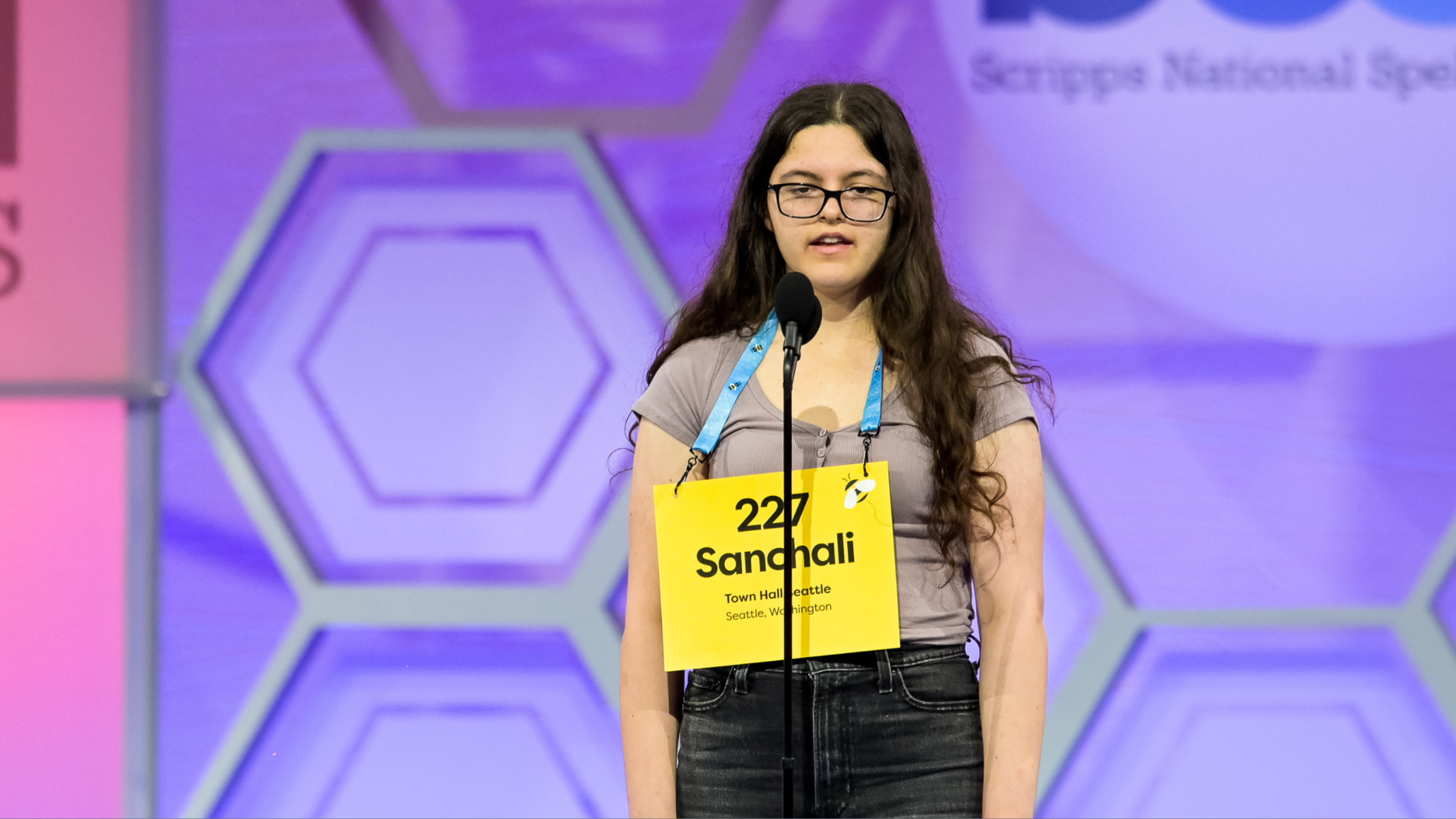 A young person with a yellow cardboard sign on their chest competes in a spelling bee