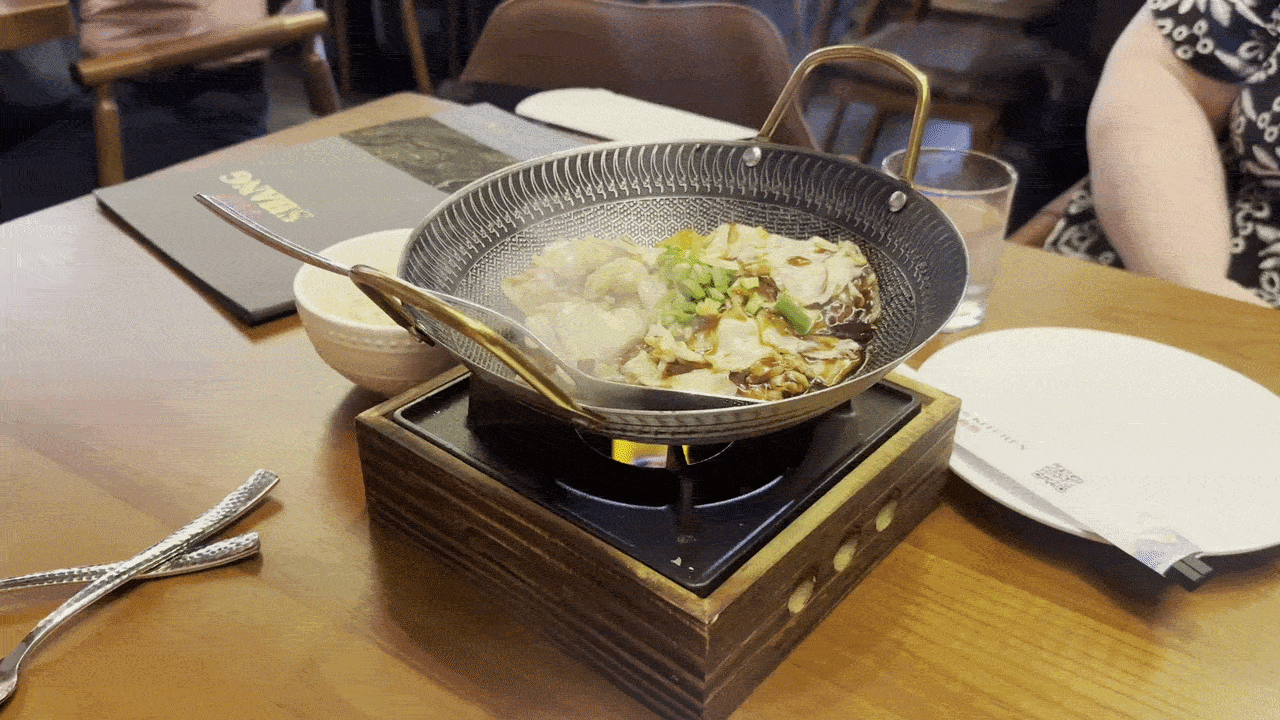 Portable stove on a wooden table heats a metal wok with a bubbling hot pot of cabbage. A spoon rests over the wok; a glass and plates sit nearby.