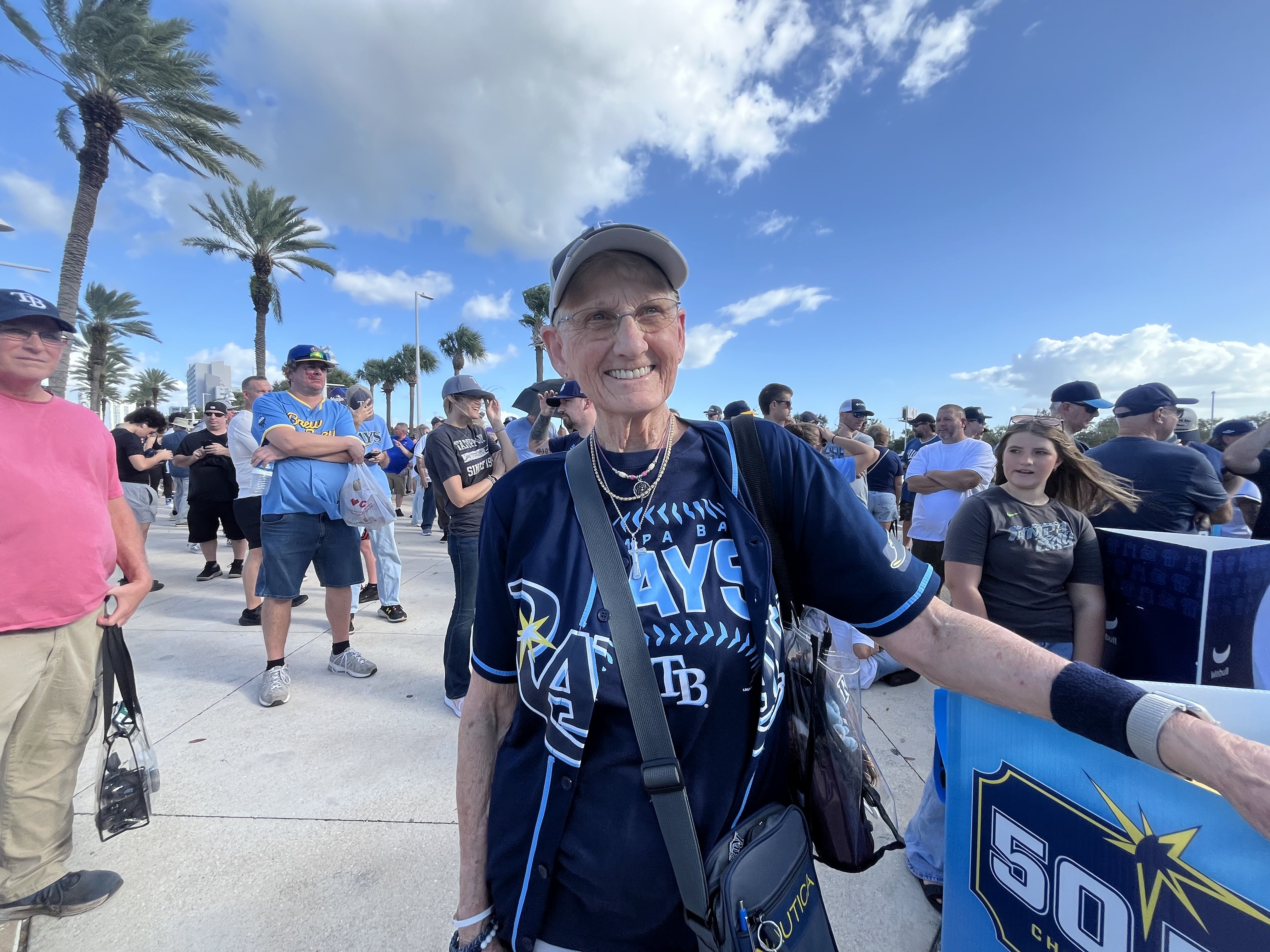 Smiling older woman wearing a cap and dark blue shirt stands at a sunny outdoor event, crowd and palm trees behind him under a bright blue sky with white clouds.
