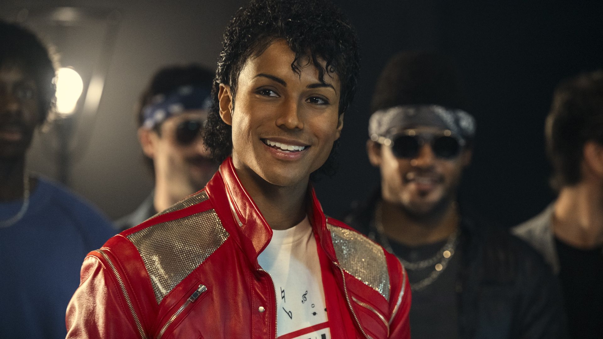 A smiling person with short curly hair wearing a red shiny jacket with silver panels over a white graphic T-shirt, in a dim backstage area with blurred people in the background.