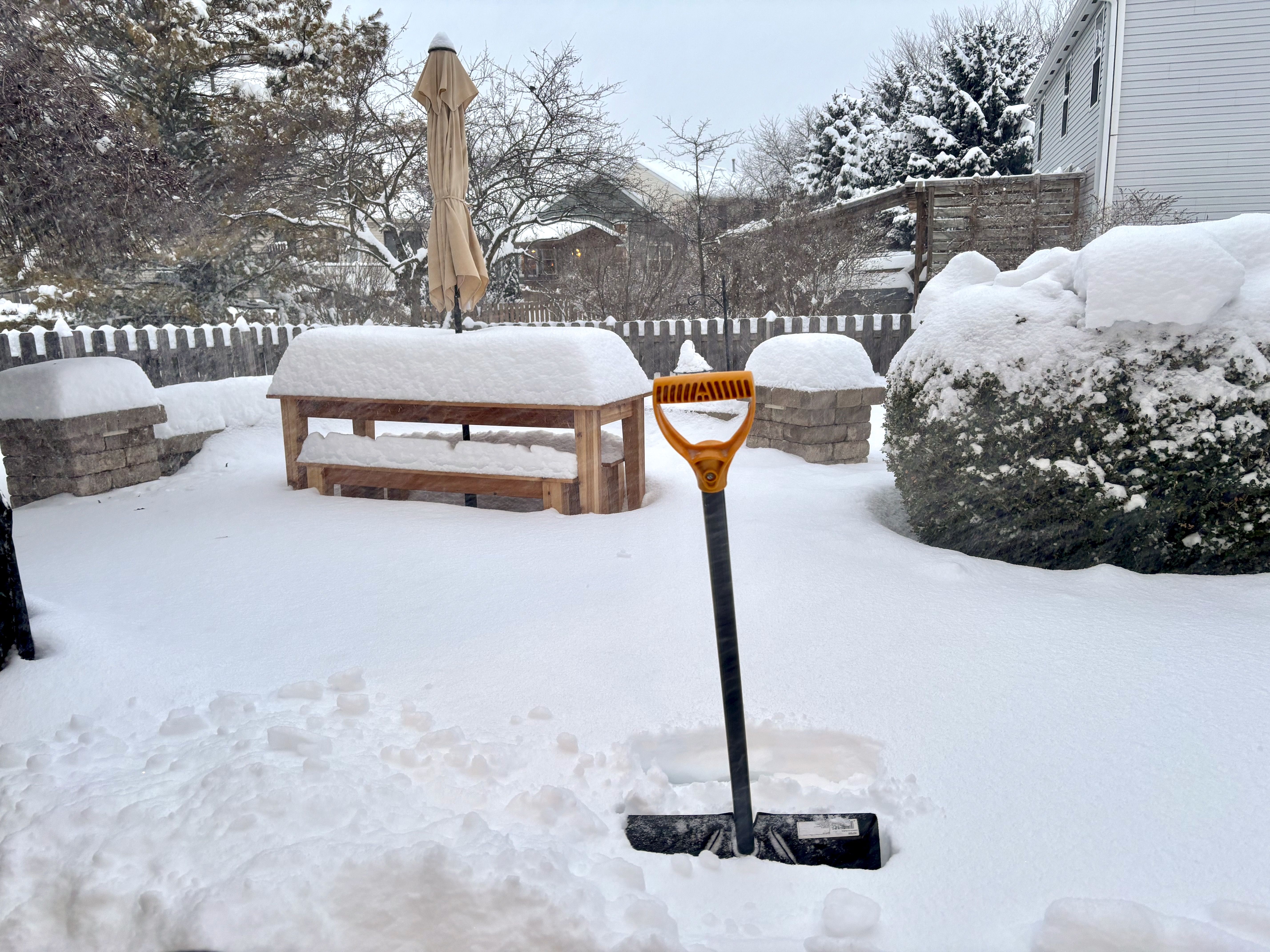 Snow-covered backyard with a wooden picnic table and benches, an umbrella, a snow shovel stuck in the snow, stone benches, and snow-covered bushes under a gray sky.