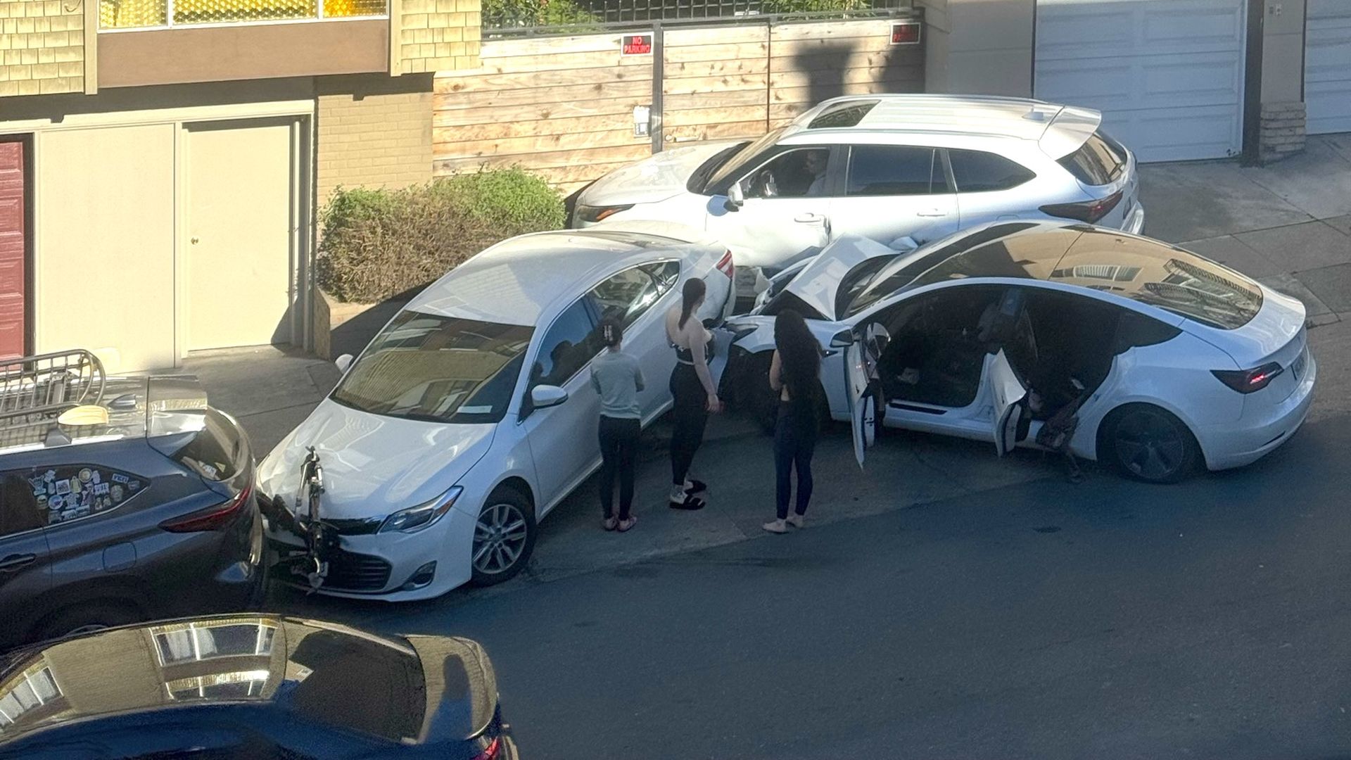Sunny urban street with beige apartment buildings; several white cars parked along the curb and a small group of people near an open car door.