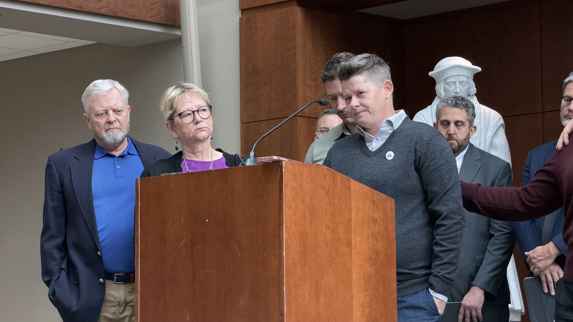 People stand near a wooden podium with a microphone as a man in a gray sweater speaks; an older man and a woman with glasses listen, with a statue bust and wood panels in the background.