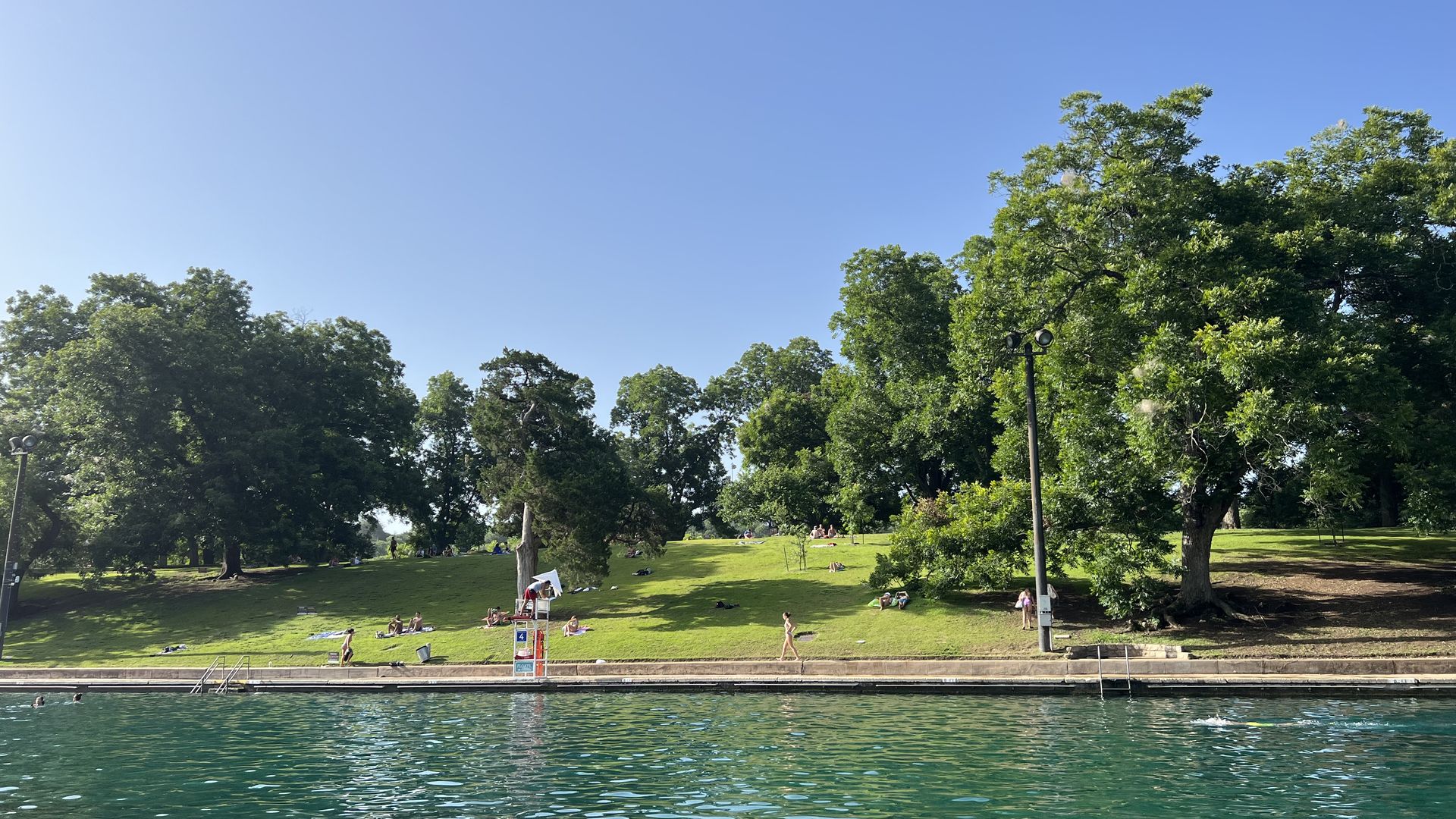 Bright, clear day at a park beside a blue lake. Green grass slopes toward the water; people lounge on blankets under leafy trees, with a lifeguard chair and distant shore.