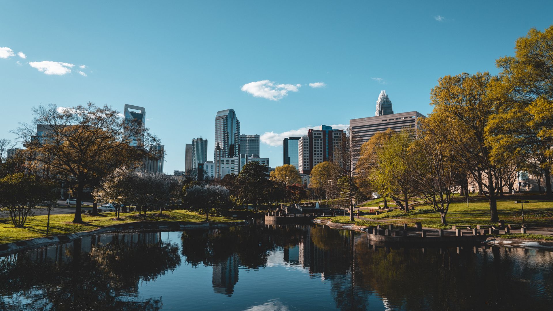 Wide city skyline under a clear blue sky with a few clouds. Modern buildings rise above a lush park by a calm river, whose surface reflects the skyline; trees show autumn yellow foliage.