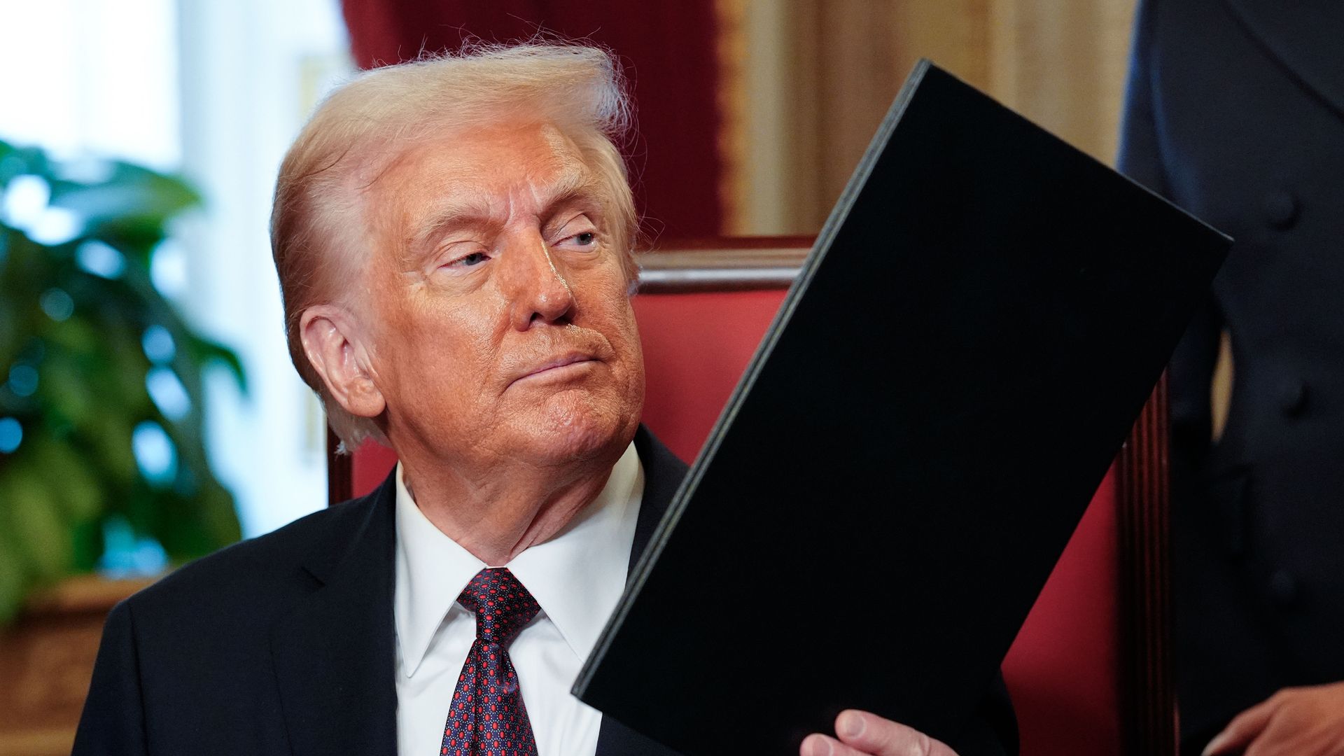 President Trump holds up a black folder after signing an executive order at his desk.