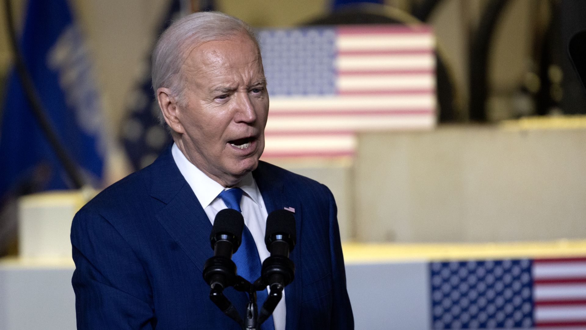 President Biden, wearing a blue suit, standing in front of American flags and speaking at microphones.