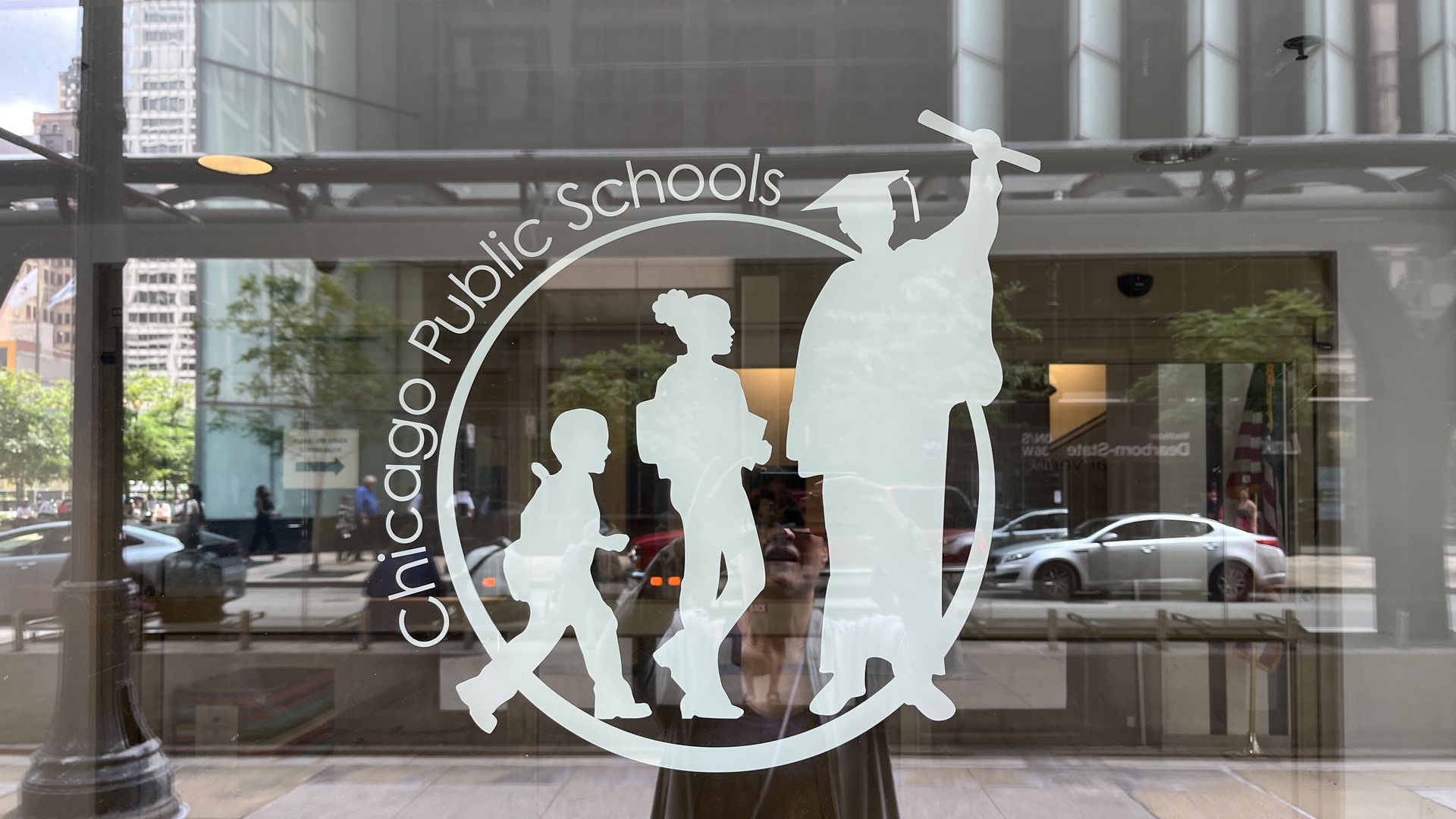 White logo on glass showing three student silhouettes progressing from young child to graduate, with text "Chicago Public Schools" and address "42 W. Madison" reflected on window.