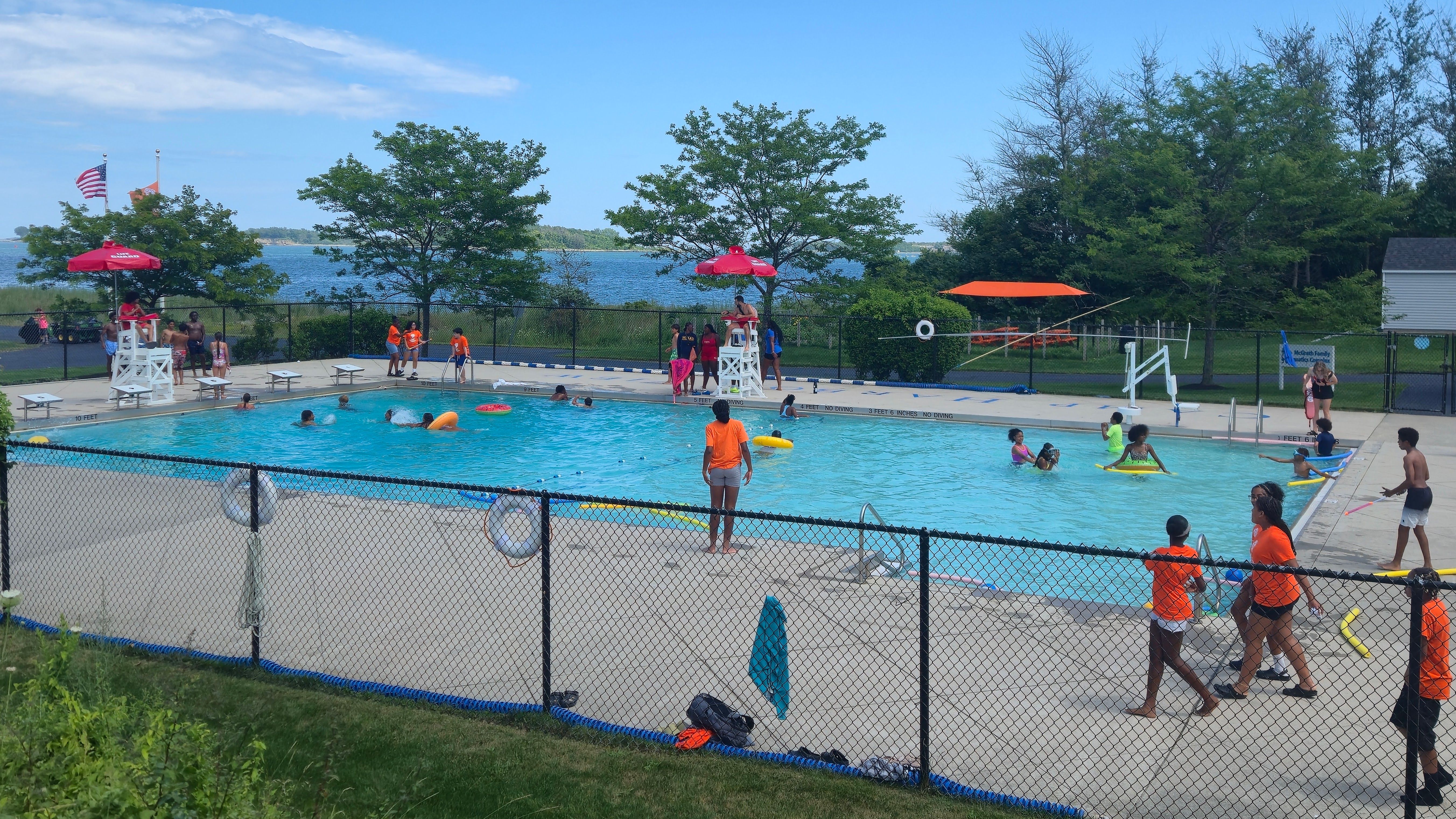 Sunny outdoor pool with kids swimming, lifeguards on white towers under red umbrellas, adults nearby. Chain-link fence, green trees, and a lake in the background; American flag nearby.