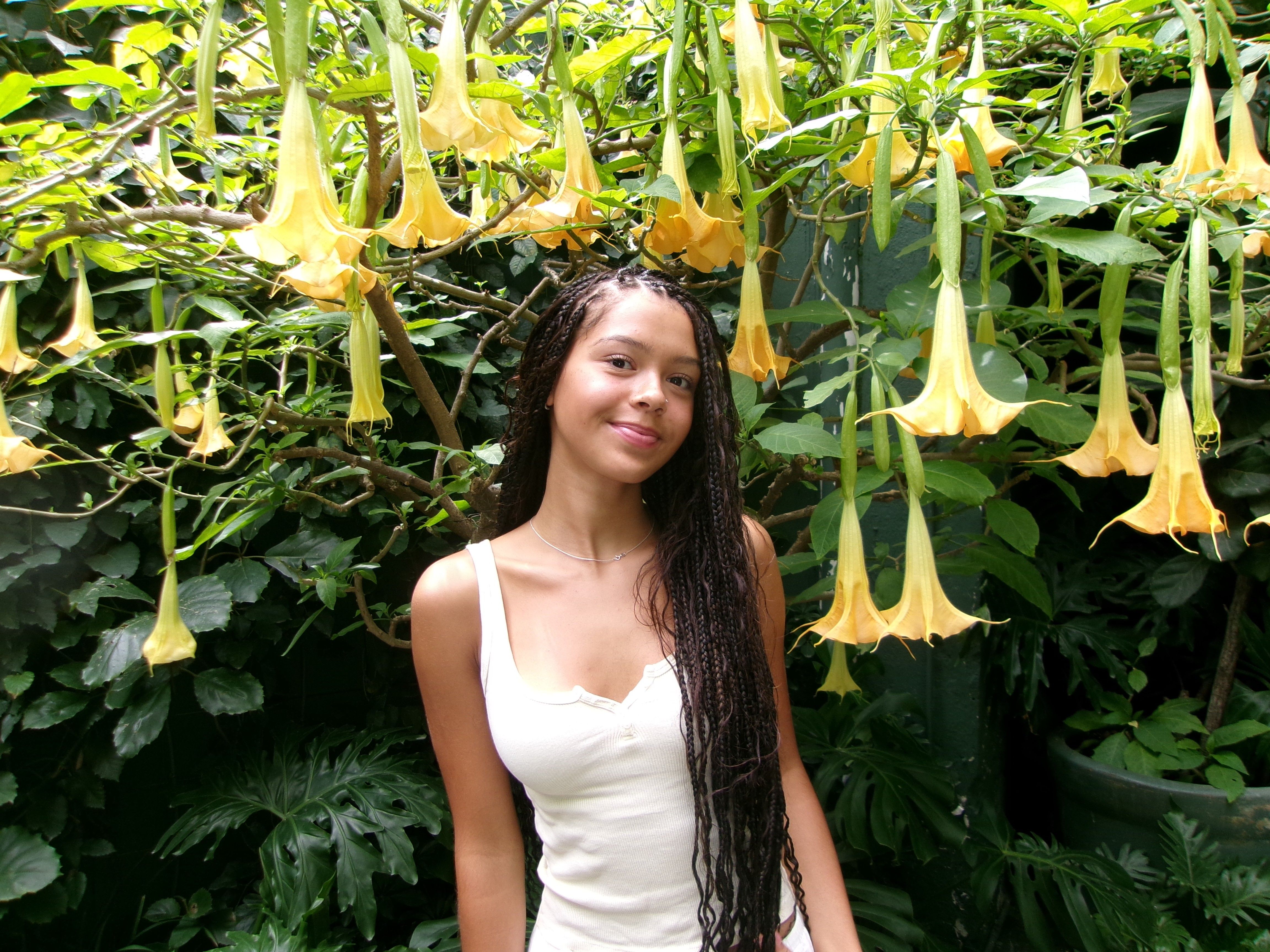 Young woman with braided hair in a white tank top stands among dense greenery and many yellow trumpet-shaped flowers.