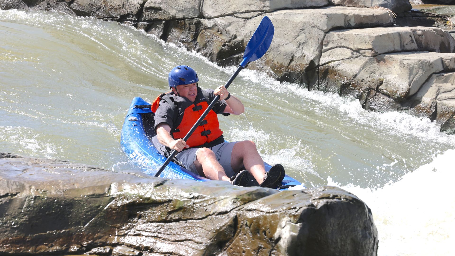A man in a blue helmet and orange life jacket paddles a blue kayak through rough whitewater, navigating between large gray rocks along a rocky riverbank.