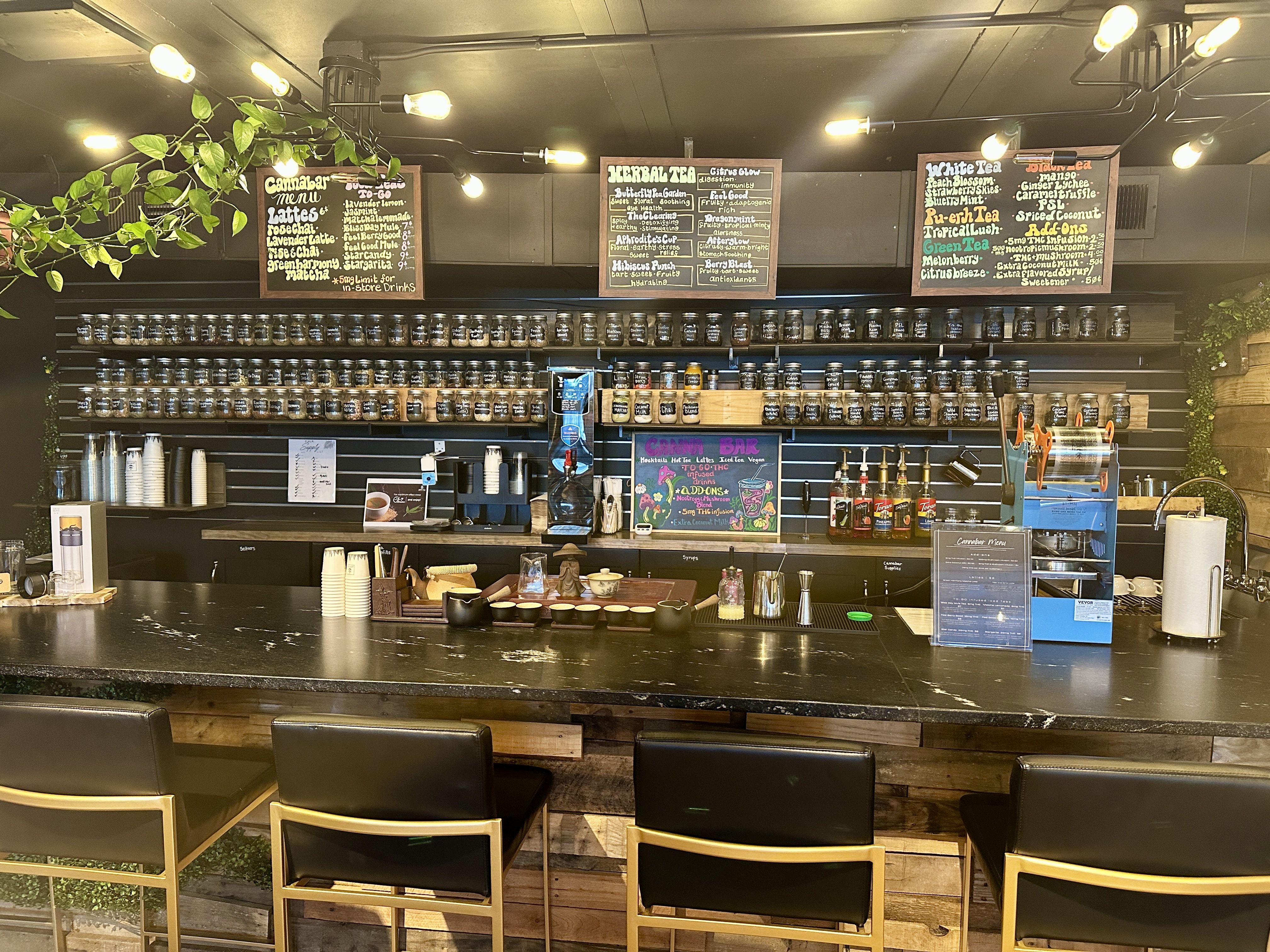 Tea shop counter with jars of tea on shelves, multiple colorful menu boards listing herbal and medicinal teas, syrups, and lattes, and four black and gold chairs at the bar.