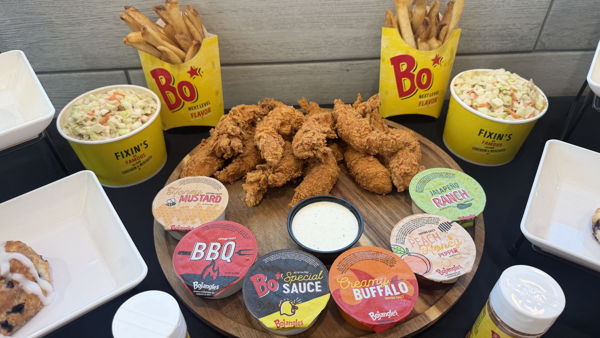 Crispy fried chicken on a wooden board, surrounded by yellow Bojangles cups filled with fries, bowls of coleslaw, and an assortment of dipping sauces labeled BBQ, Buffalo, and honey mustard.