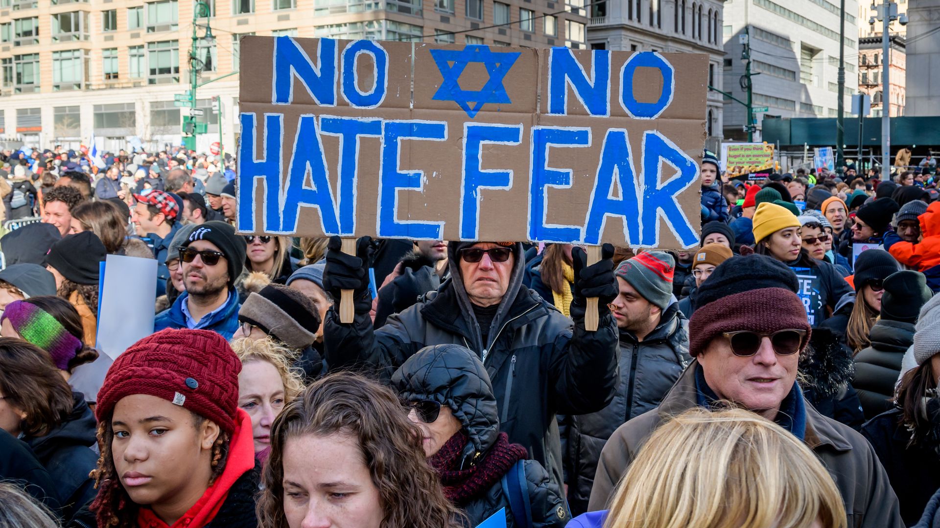 Photo of a crowd of people with one holding a cardboard sign with blue lettering that says "No hate, no fear" and shows the Star of David