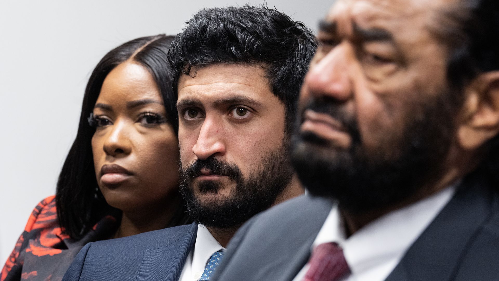 Three serious adults in business attire seated closely, with a woman in a red and black patterned outfit on the left, a man in a blue suit and patterned tie in the center, and an older man in a suit and red tie on the right.