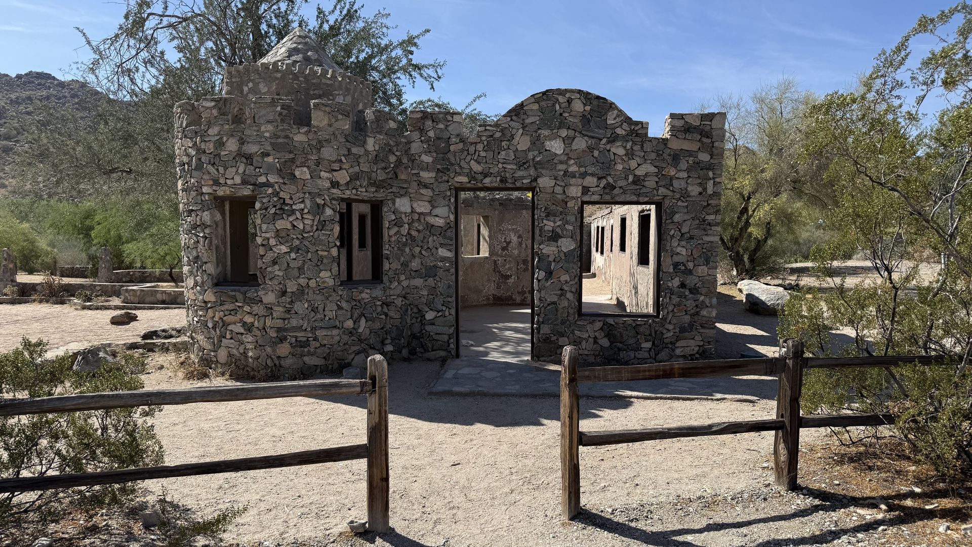 Faux desert stone ruin with a round-topped turret, thick stone walls and multiple door and window openings. Dry brush and shrubs surround the structure under a clear blue sky, with a wooden fence in front.