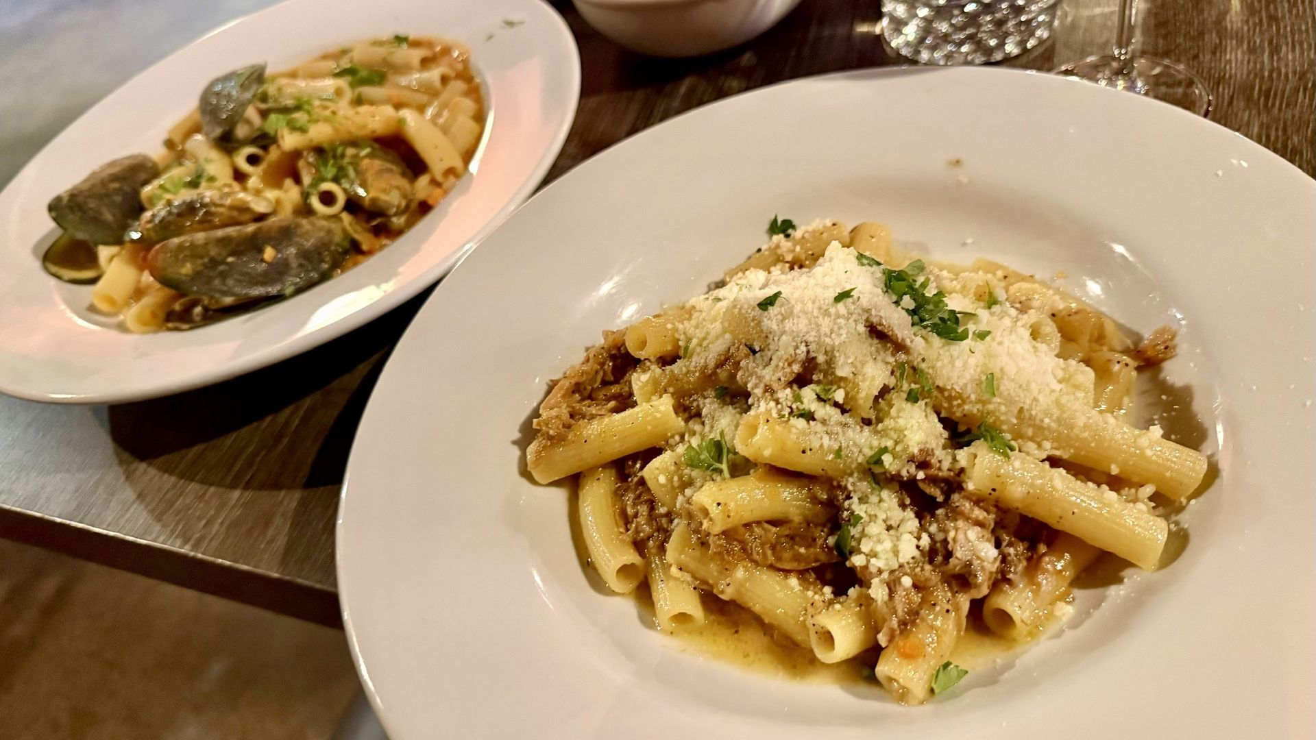 Two plates of pasta on a wooden table: foreground rigatoni with grated cheese and parsley; background mussels pasta. Bread basket, water glass, and a wine glass nearby.