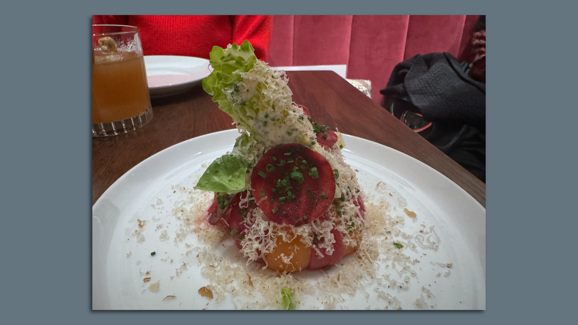 A close-up image of a plate with a small pile of beets, upon which is a small piece of lettuce sticking out of the top. It's all covered in shaved cheese and green onions.