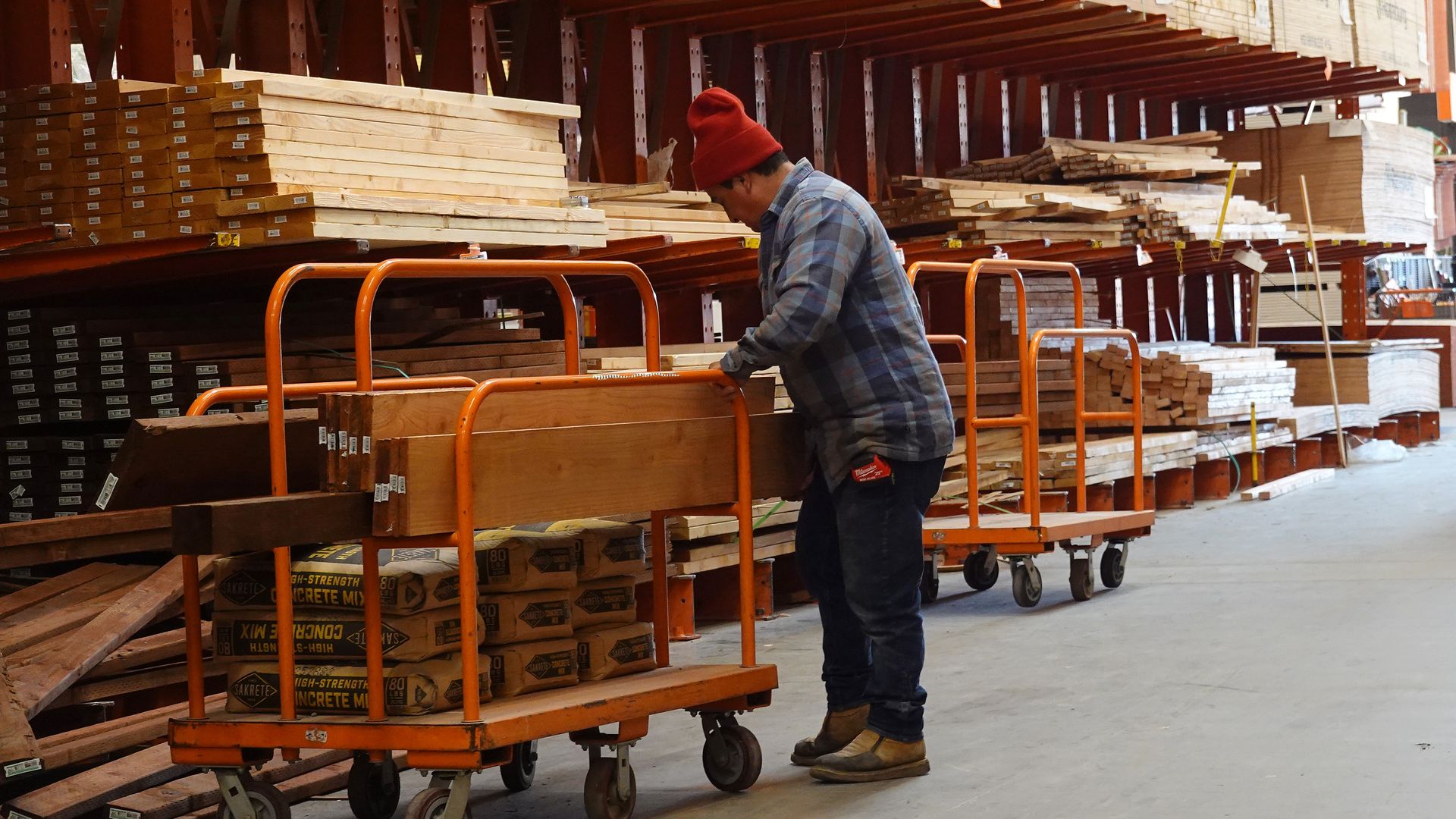 A person wearing a red beanie and plaid shirt loading wood onto an orange cart in a warehouse with stacks of lumber and bags of concrete mix.