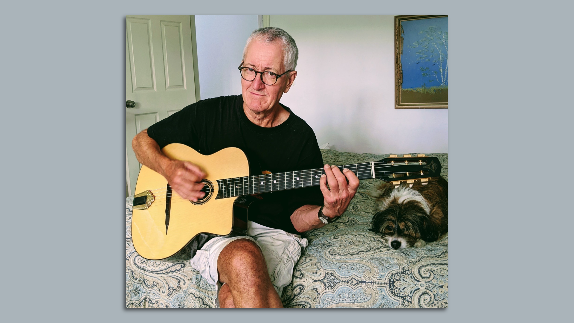 Photo of a man sitting on a bed with a dog. The man is holding a guitar. 