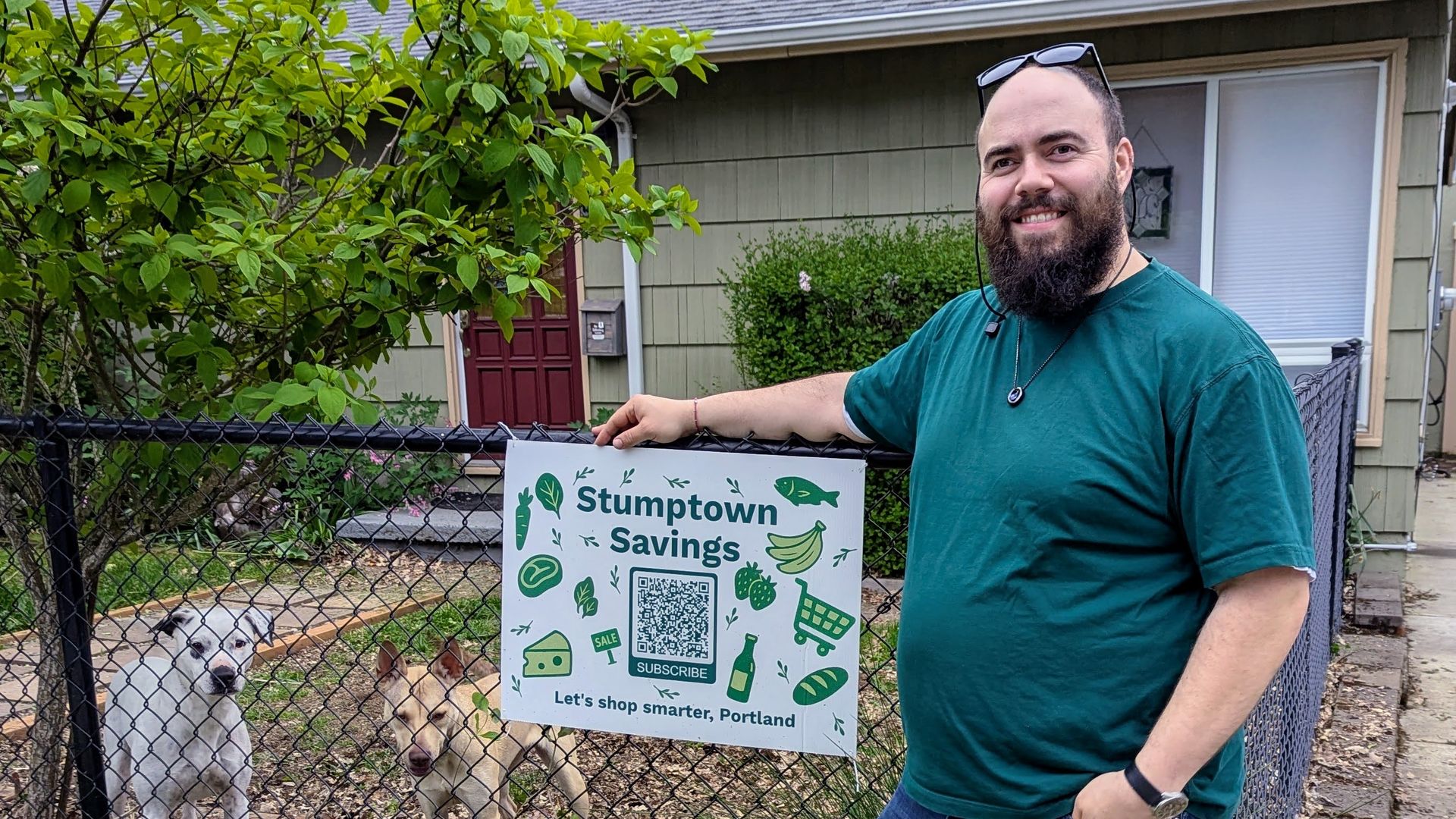 A man stands next to a fence with a sign advertising his newsletter called Stumptown Savings with two cute dogs looking on from behind a fence.