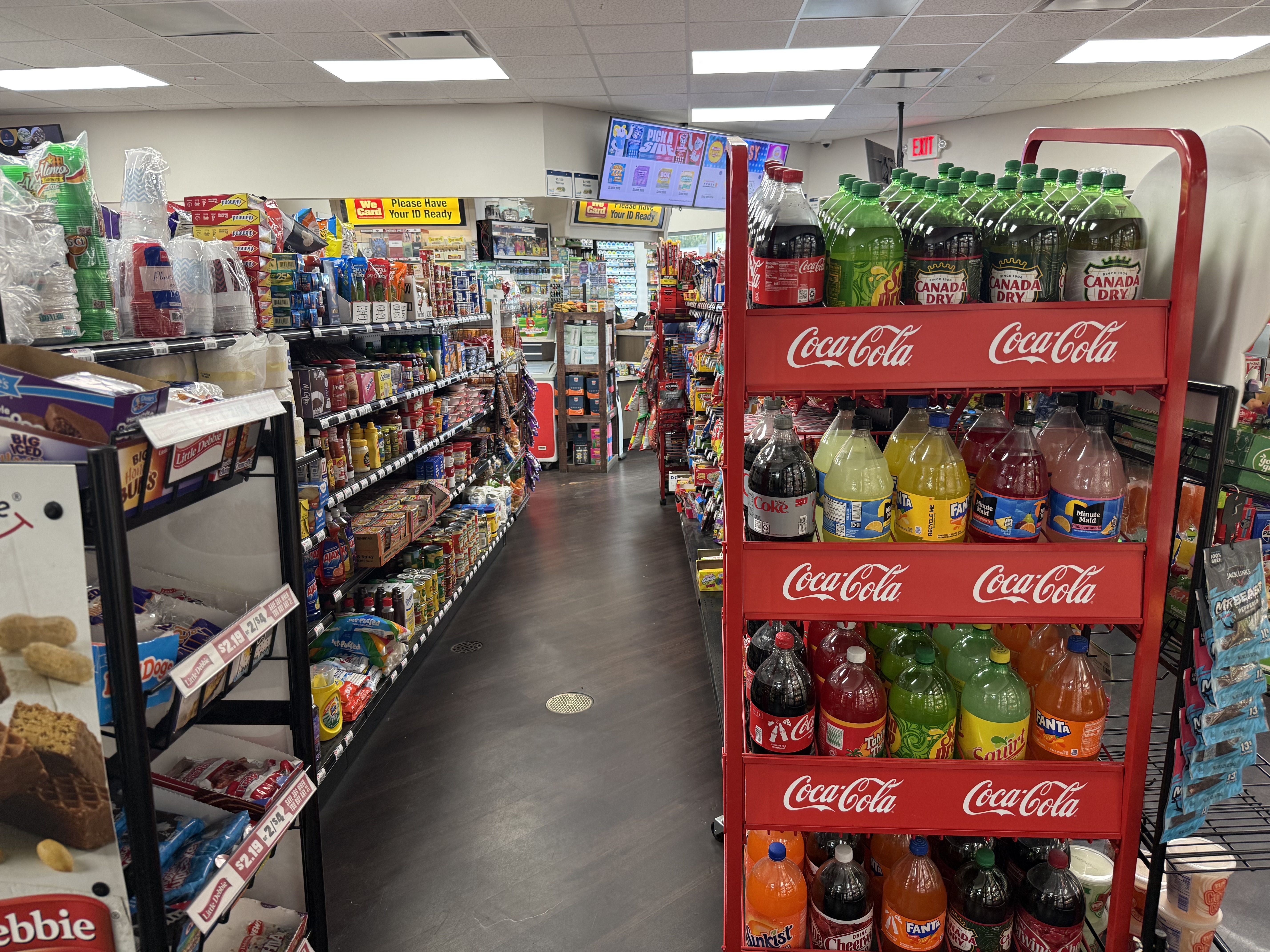 Inside a convenience store, long aisles are stocked with snacks and drinks. A bright red Coca‑Cola display rack on the right holds many large bottles, with a back checkout area and overhead digital menu behind.