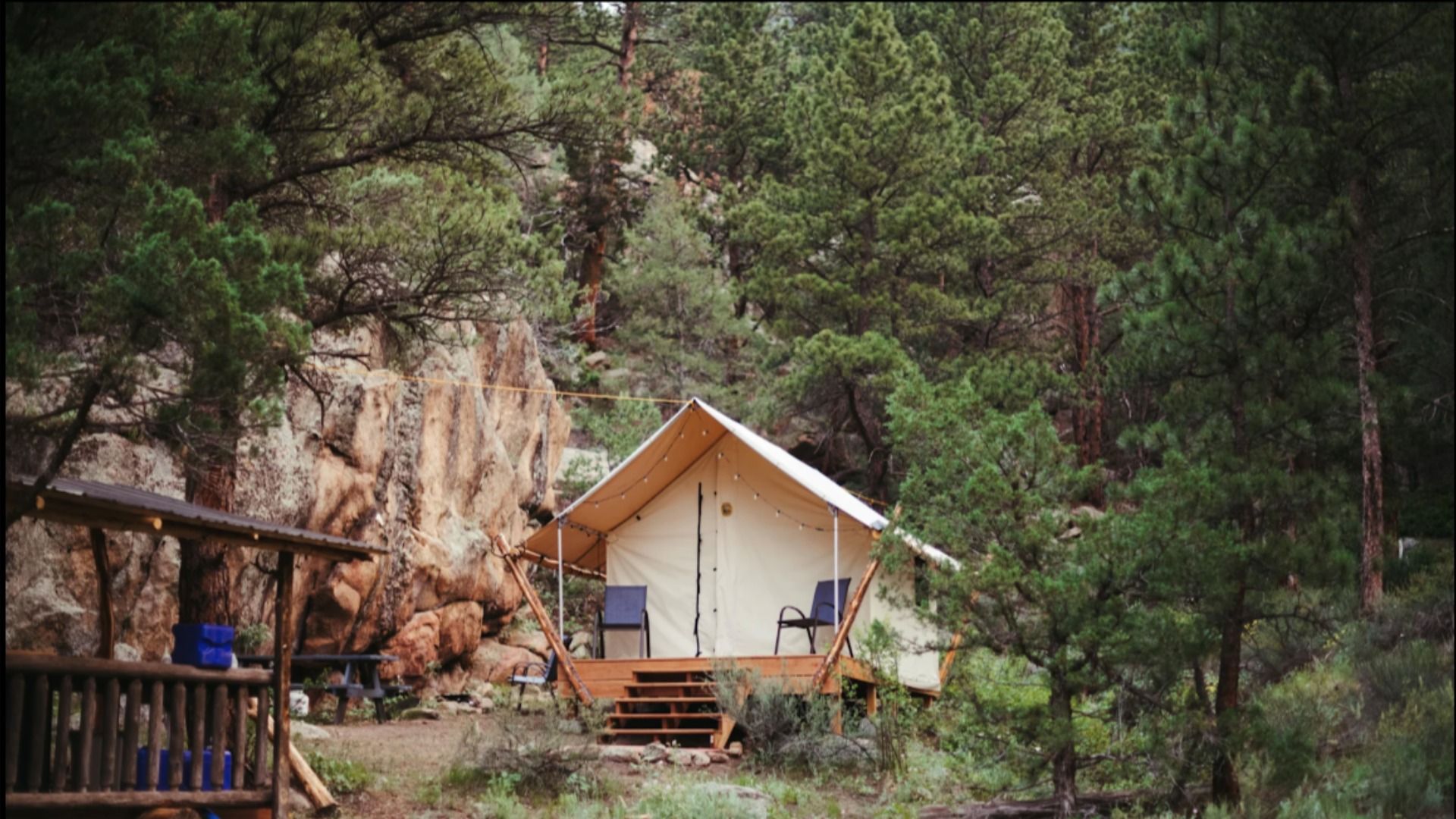 Canvas tent with two chairs on wooden porch in a forest clearing surrounded by pine trees and large rocks, with a rustic wooden shelter to the left.