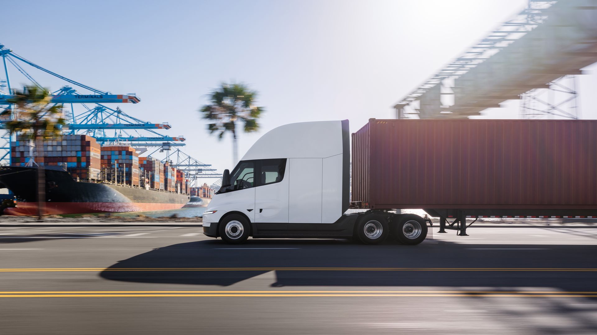 A white semi-truck with a brown shipping-container trailer drives along a port road; behind it, colorful stacked containers and blue gantry cranes rise against a clear blue sky.