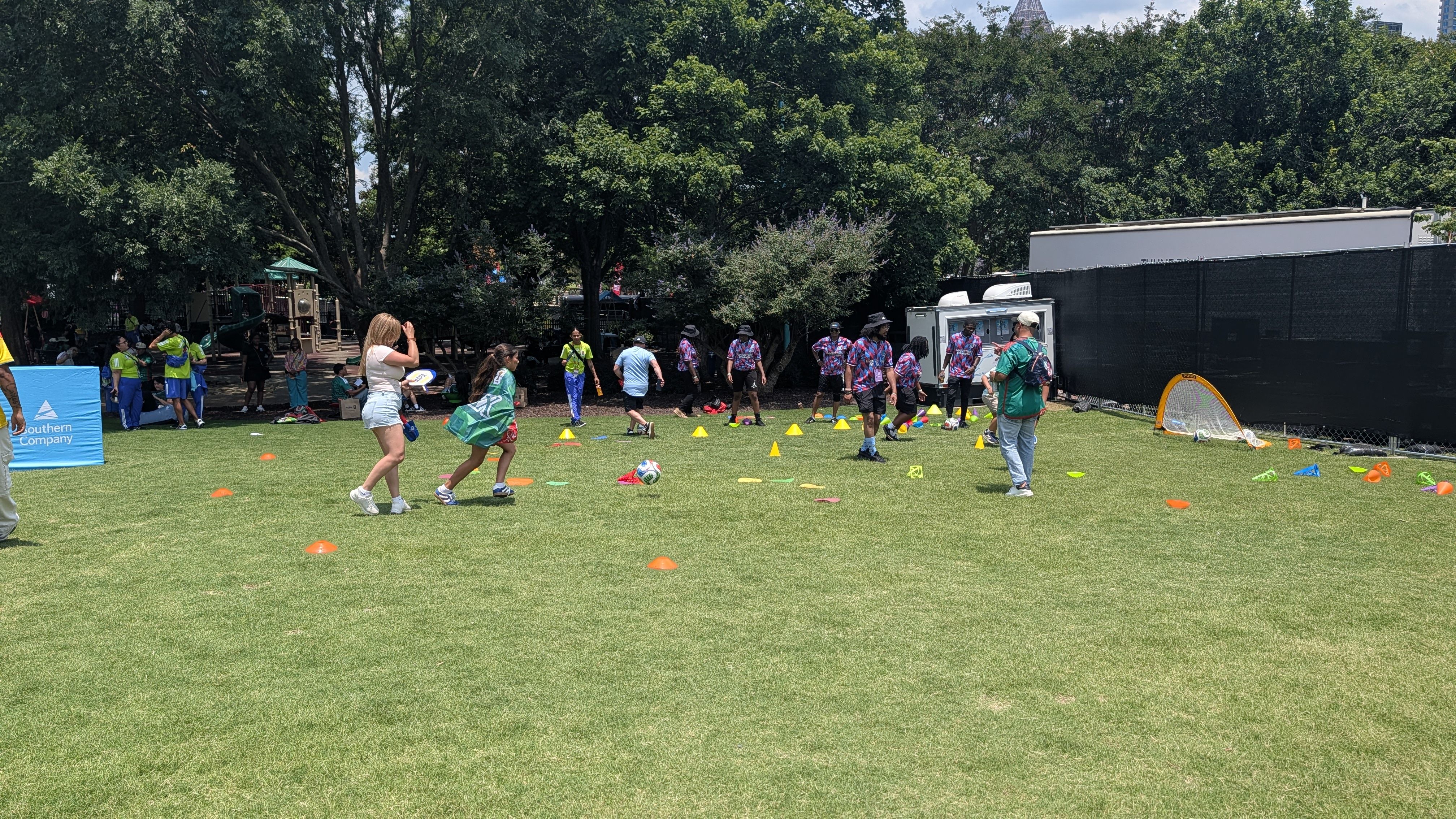 A sunny park scene with kids and adults on a grassy field, cones set for a game, players in bright shirts, a small goal, a blue canopy, and tall trees behind a black fence.