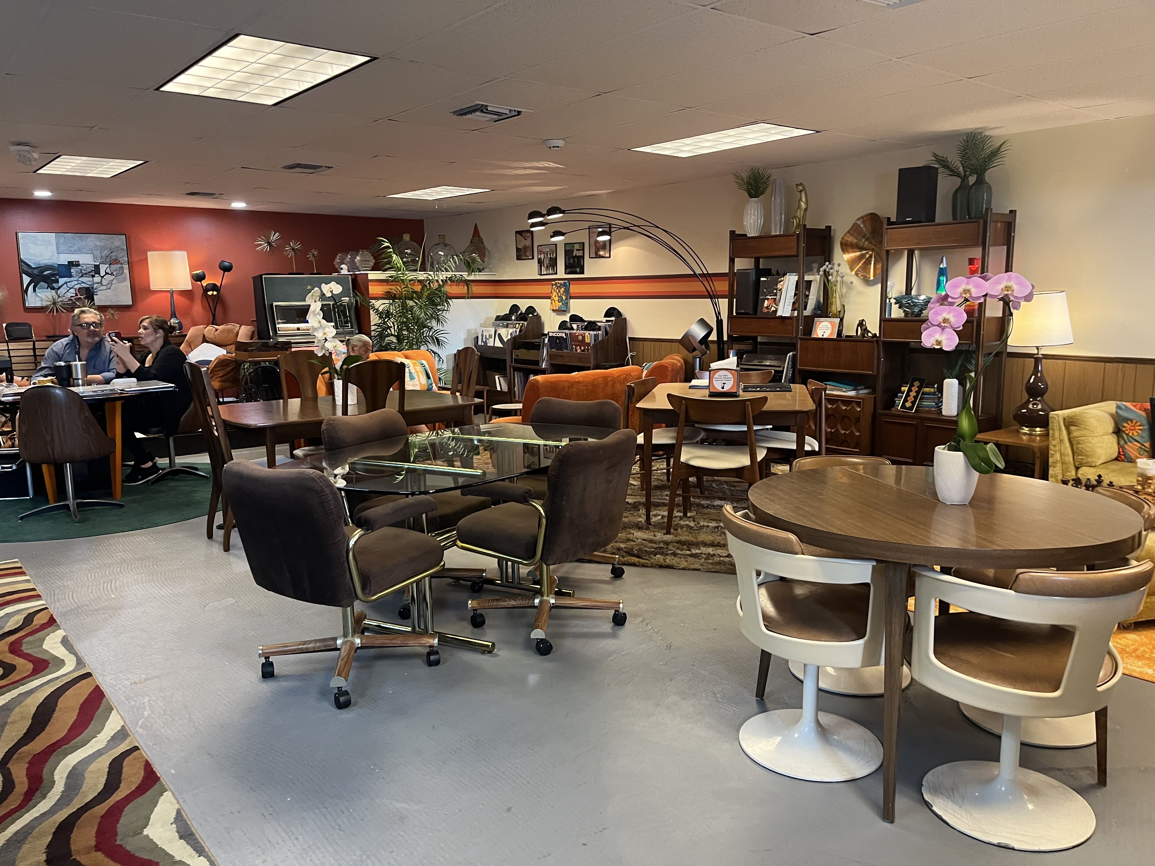 Inside a retro-style lounge with many wooden tables and chairs. A red wall on the left, potted plants and wall art; foreground pink orchid. People sit at a table; tall shelves with decor on the right.