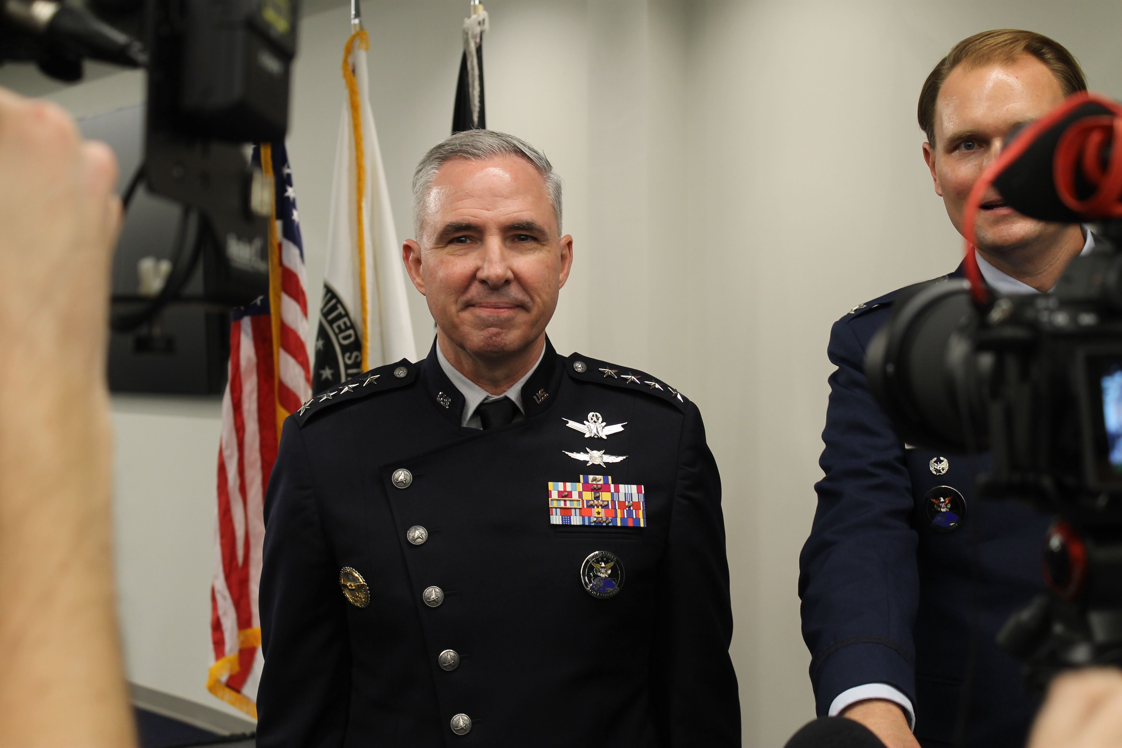 Center: a senior police officer in dress uniform with ribbons and badges, standing before flags. A microphone blocks part of another officer on the right; photographer's hand in foreground.