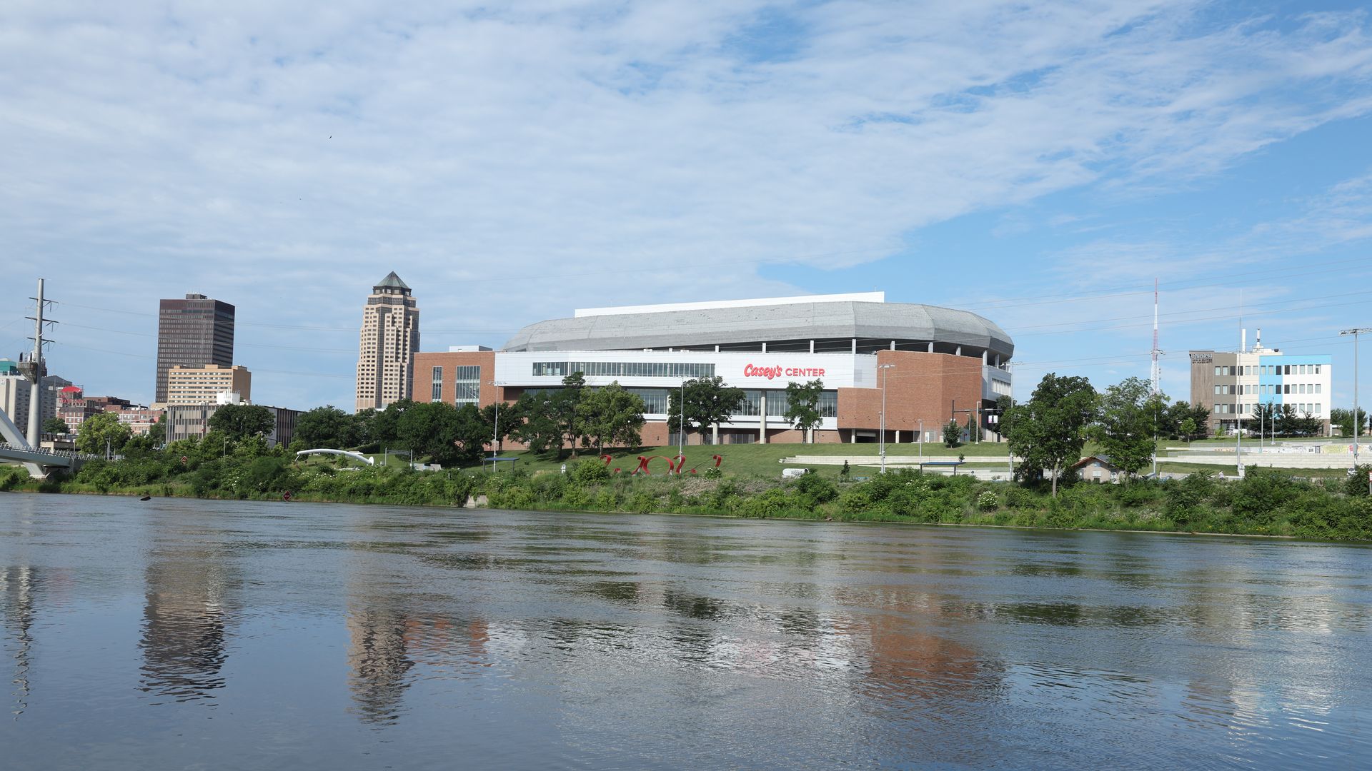 Wide river in the foreground with green banks, a modern stadium labeled "Casey's Center" in the midground, brick buildings and a skyline of tall office towers under a clear blue sky.