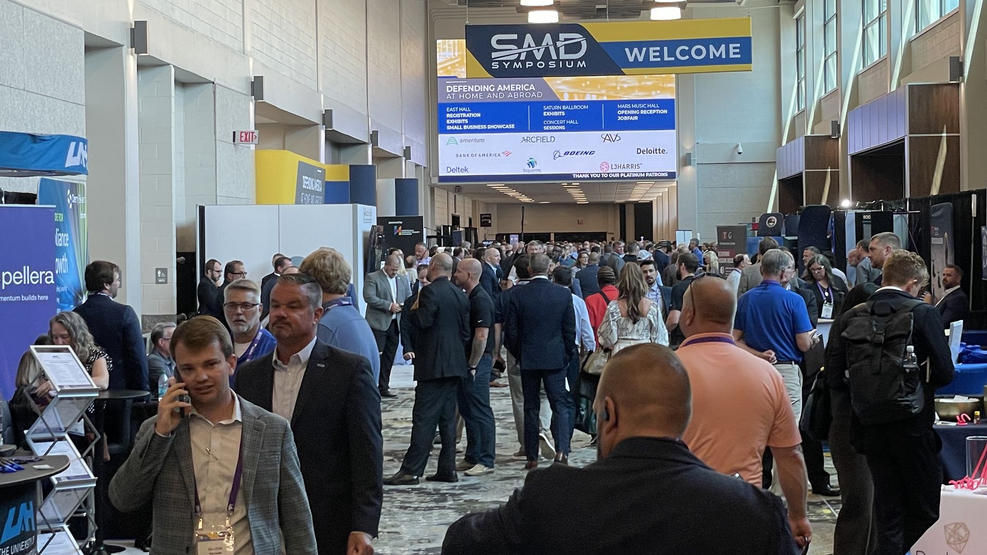 Busy conference hall with a large blue-yellow SAMD Symposium banner and welcome sign. Crowds of professionals mingle between exhibitor booths, white walls, and tall windows along the side.