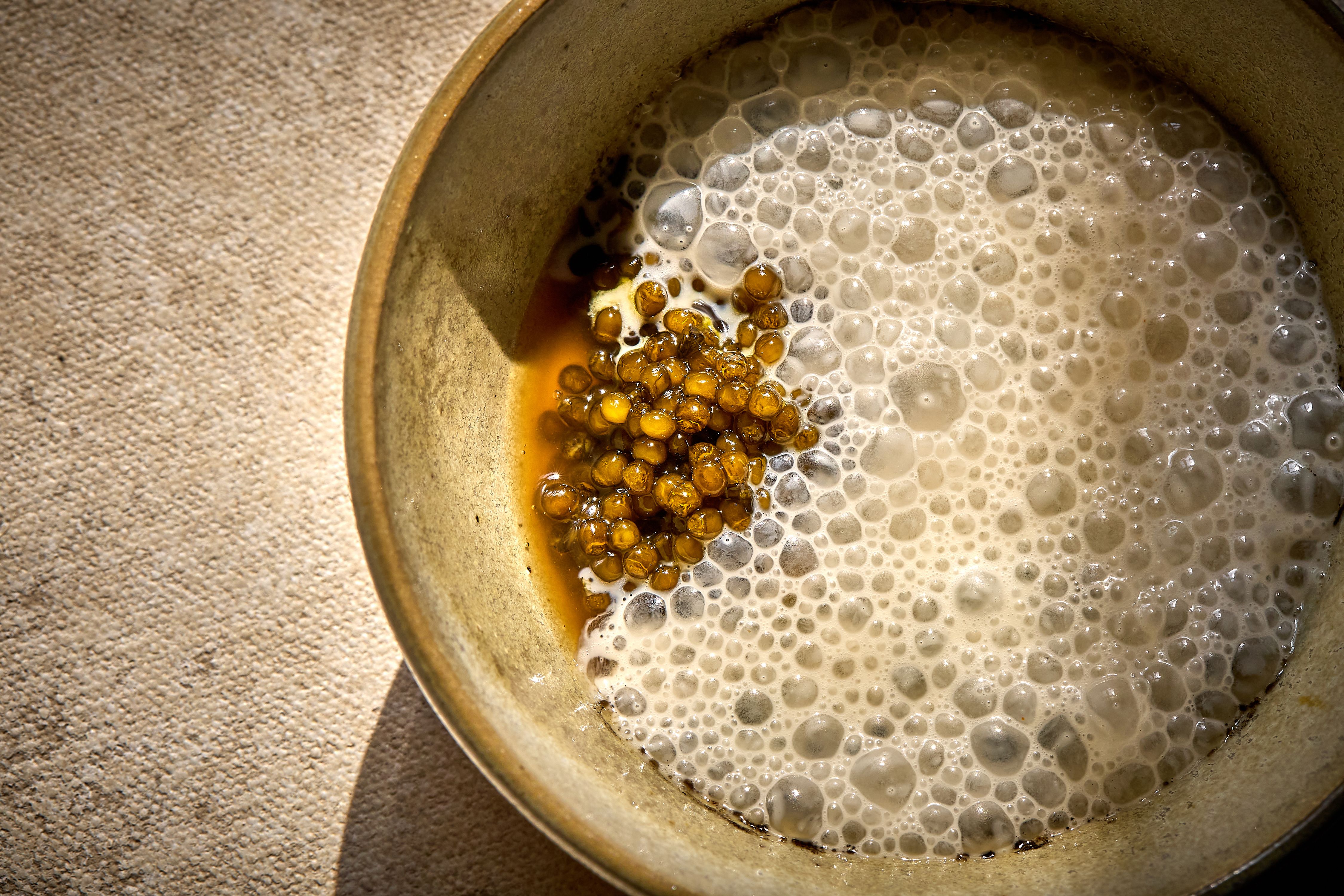 Close-up of a beige ceramic bowl containing a foamy, light brown liquid and a cluster of small, translucent amber-colored bubbles on a textured beige surface.