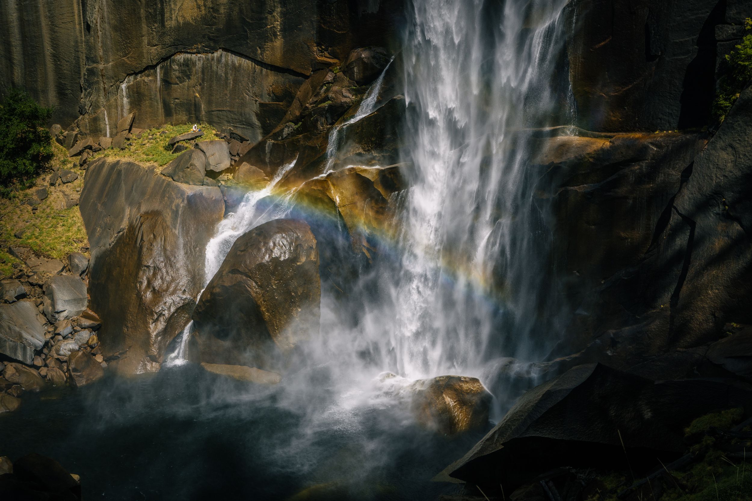 Waterfall cascading over large rocks with mist and a rainbow forming in the spray, surrounded by rugged, dark brown and green rocky cliffs in sunlight.