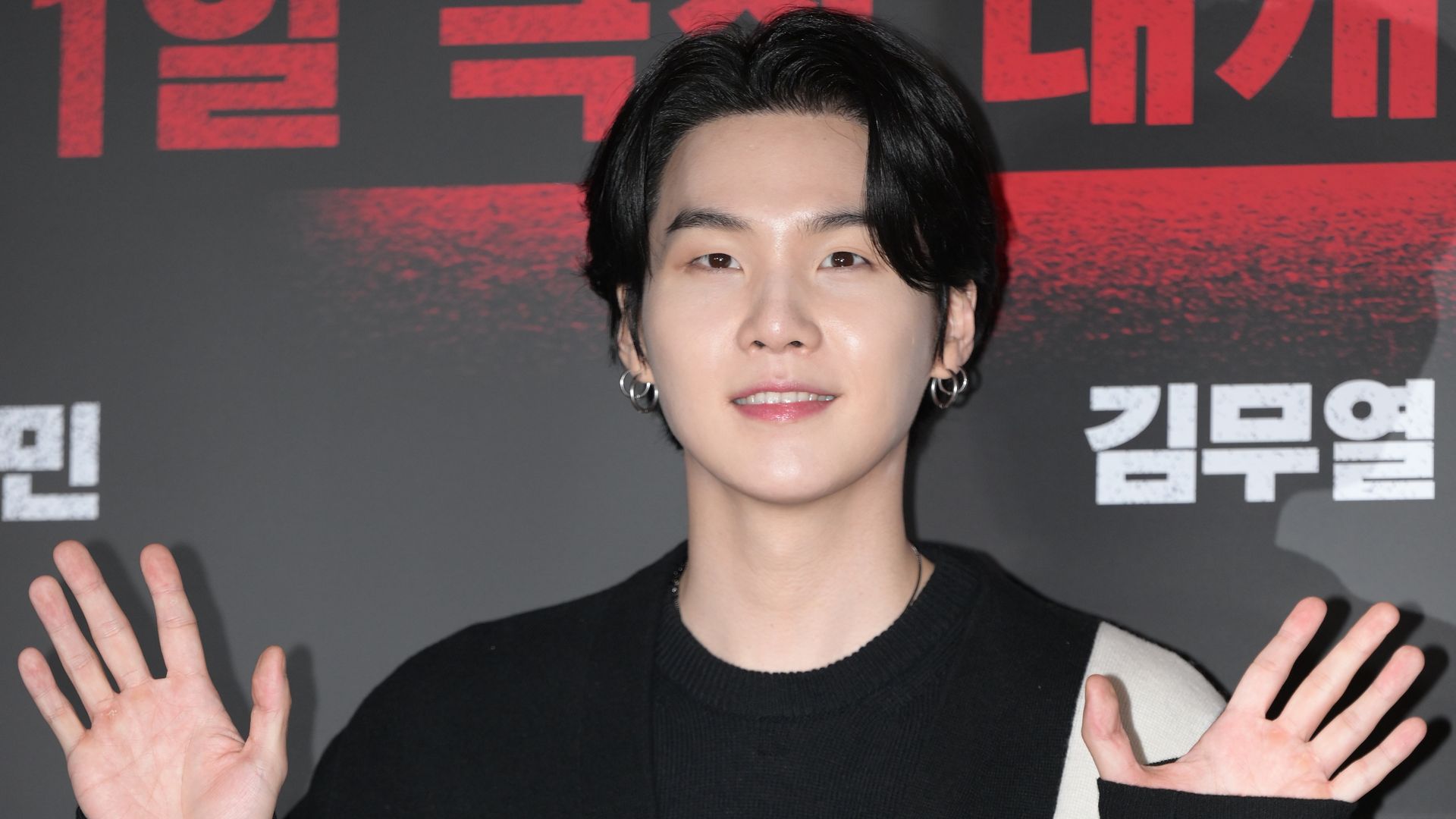 Person with short black hair and silver hoop earrings, wearing a black sweater, smiling at the camera with hands raised. A red-and-black backdrop with large block letters.