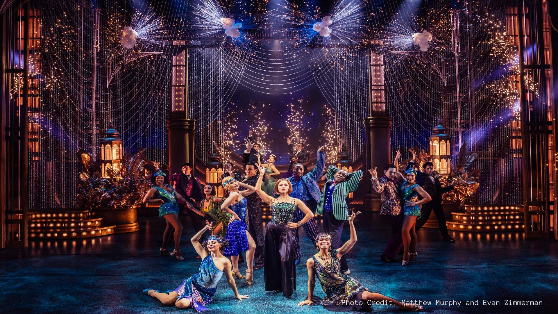  A vibrant musical cast poses mid-performance in front of a glittering, firework-lit stage set in a glamorous, Gatsby-inspired scene.