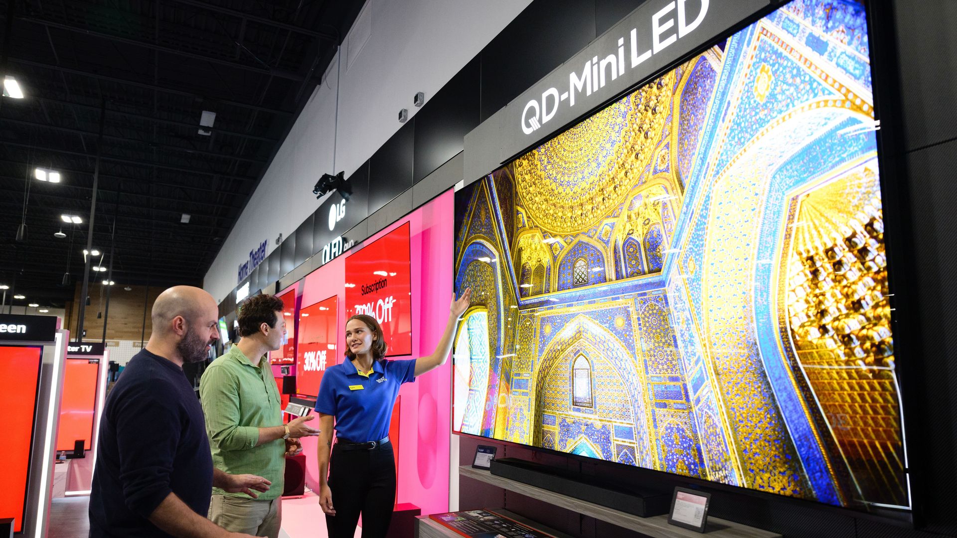A Best Buy store employee in a blue shirt shows two customers a large QD-Mini LED TV displaying a massive television.