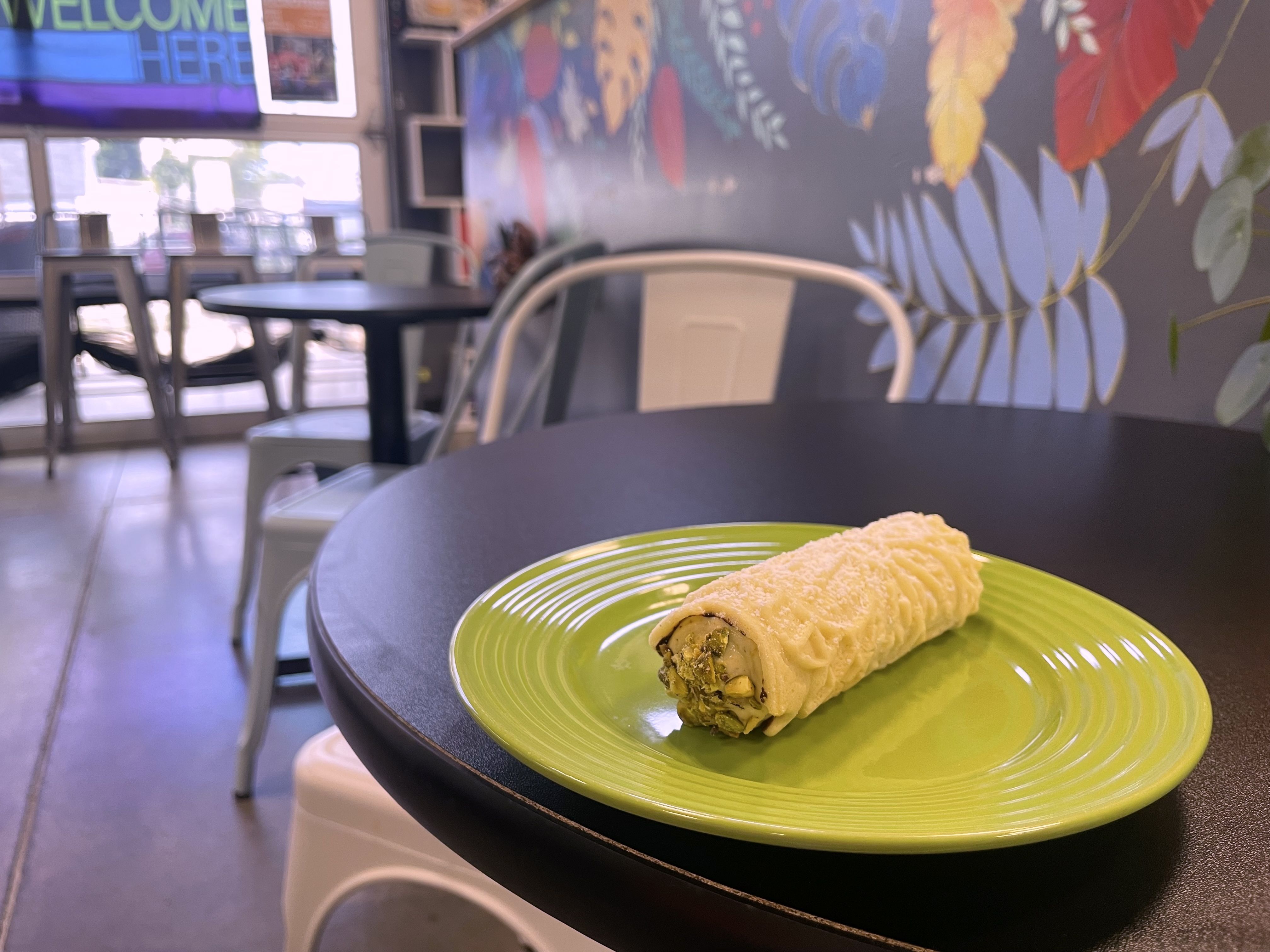 Bright green plate with a rolled pastry on a dark cafe table in the foreground; blurred background shows tables, chairs, and a colorful leaf mural on the wall.
