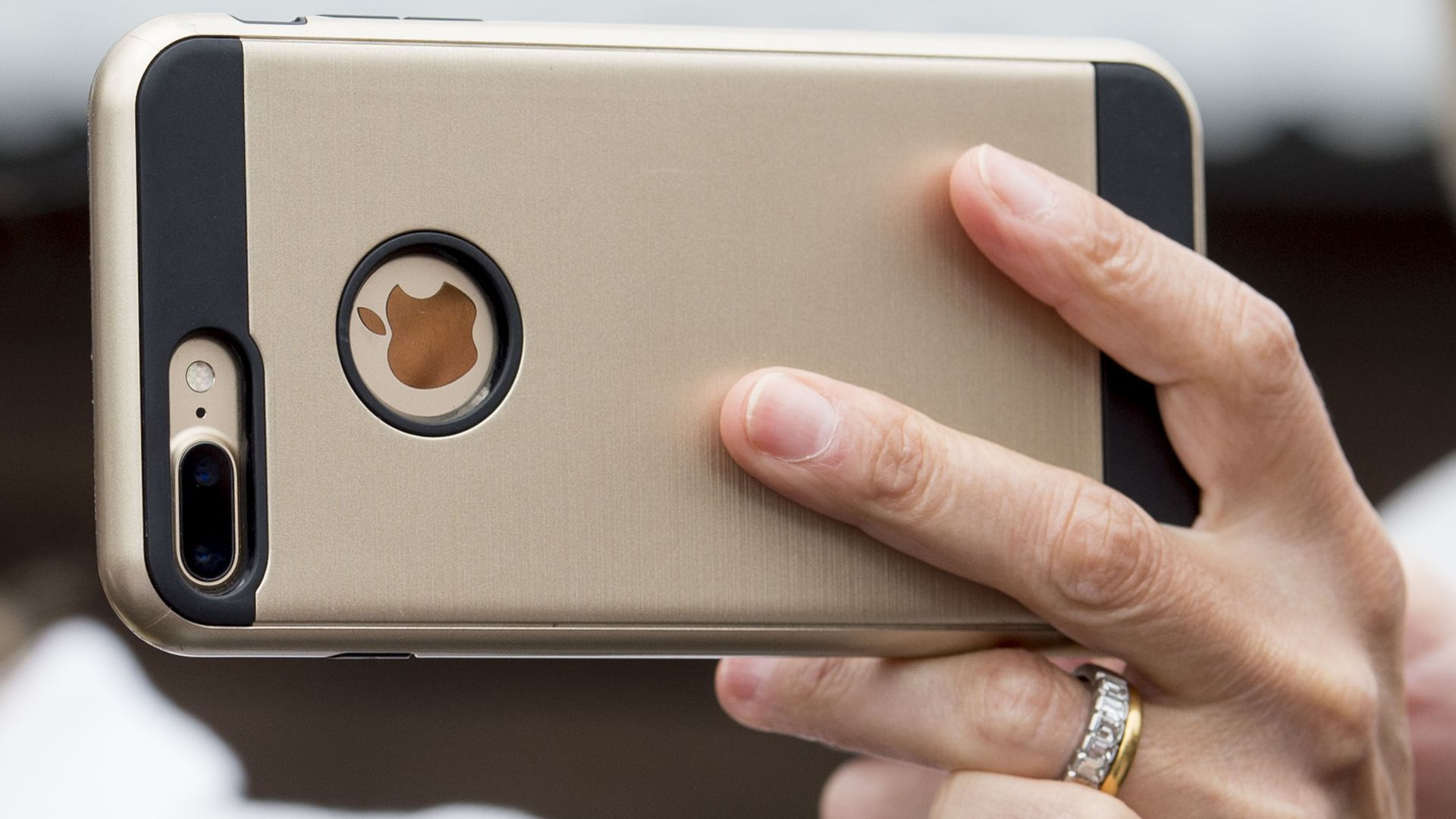 Close-up of a gold iPhone held by two hands; visible camera module on the left, circular Apple logo ring, black bumper, beige back, blurred background.