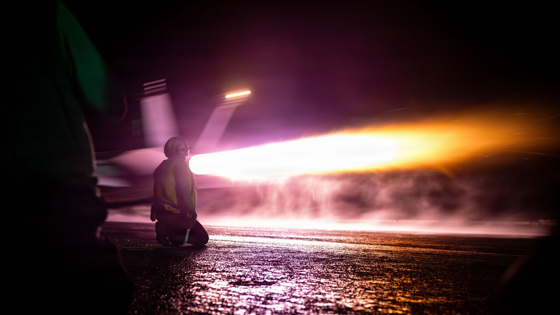 Person in safety gear kneeling near an aircraft jet engine emitting bright orange and white flames at night on tarmac.